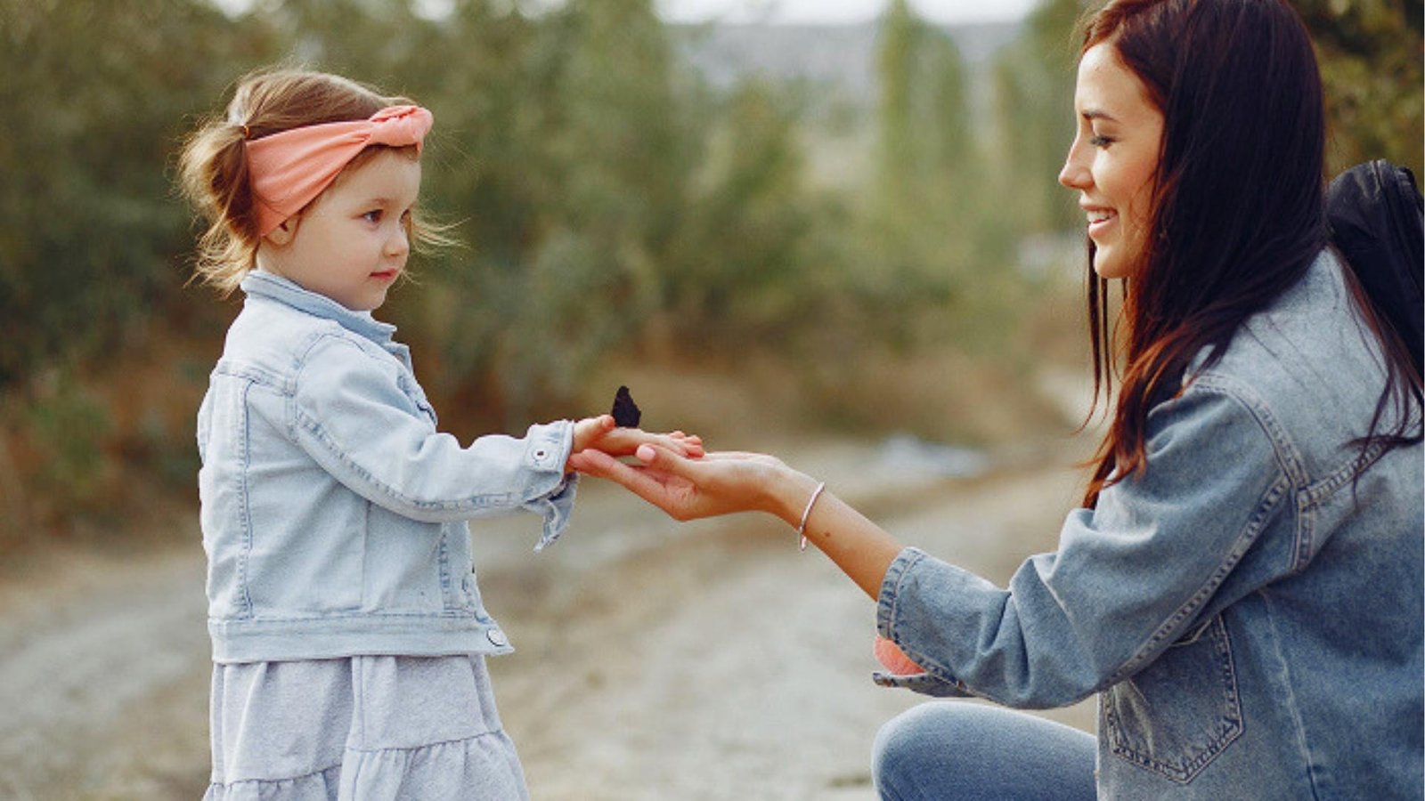 Young girl in denim and headband offers butterfly on her palm to kneeling woman outdoors, gentle mindful moment.