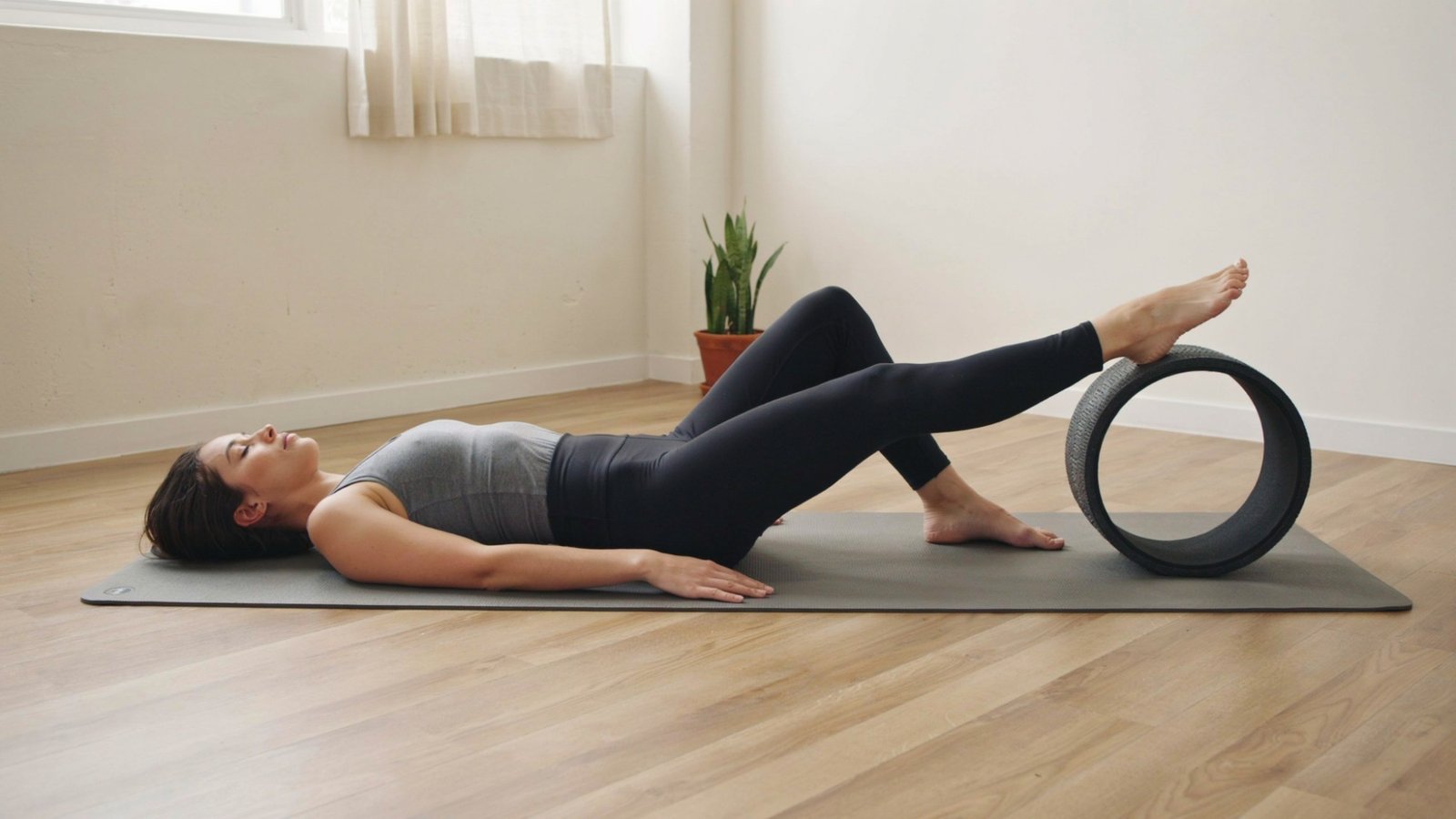 Person lies on a yoga mat with one foot resting on the yoga wheel for support.