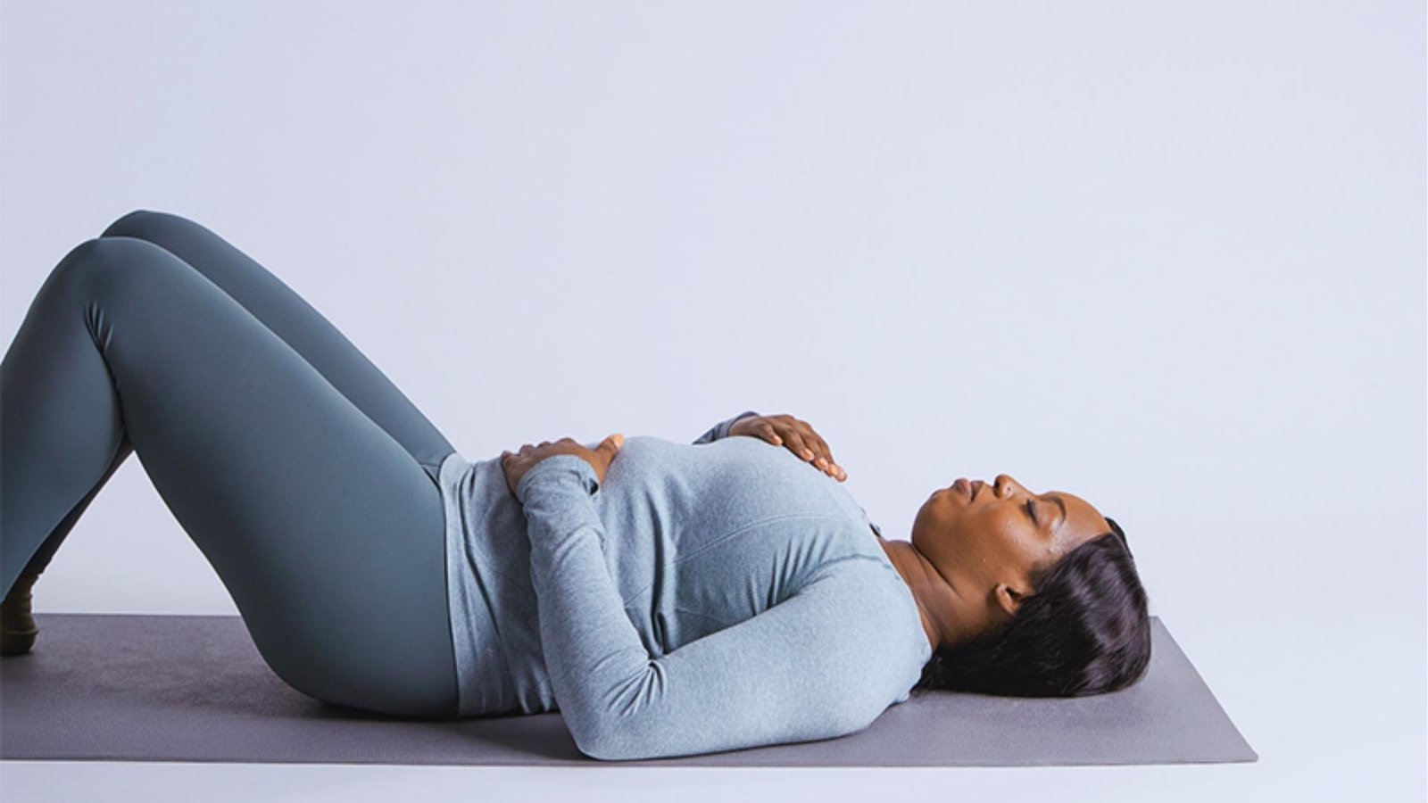 Woman in gray top and leggings lies on mat with hands on ribs, practicing diaphragmatic breathing for core engagement.