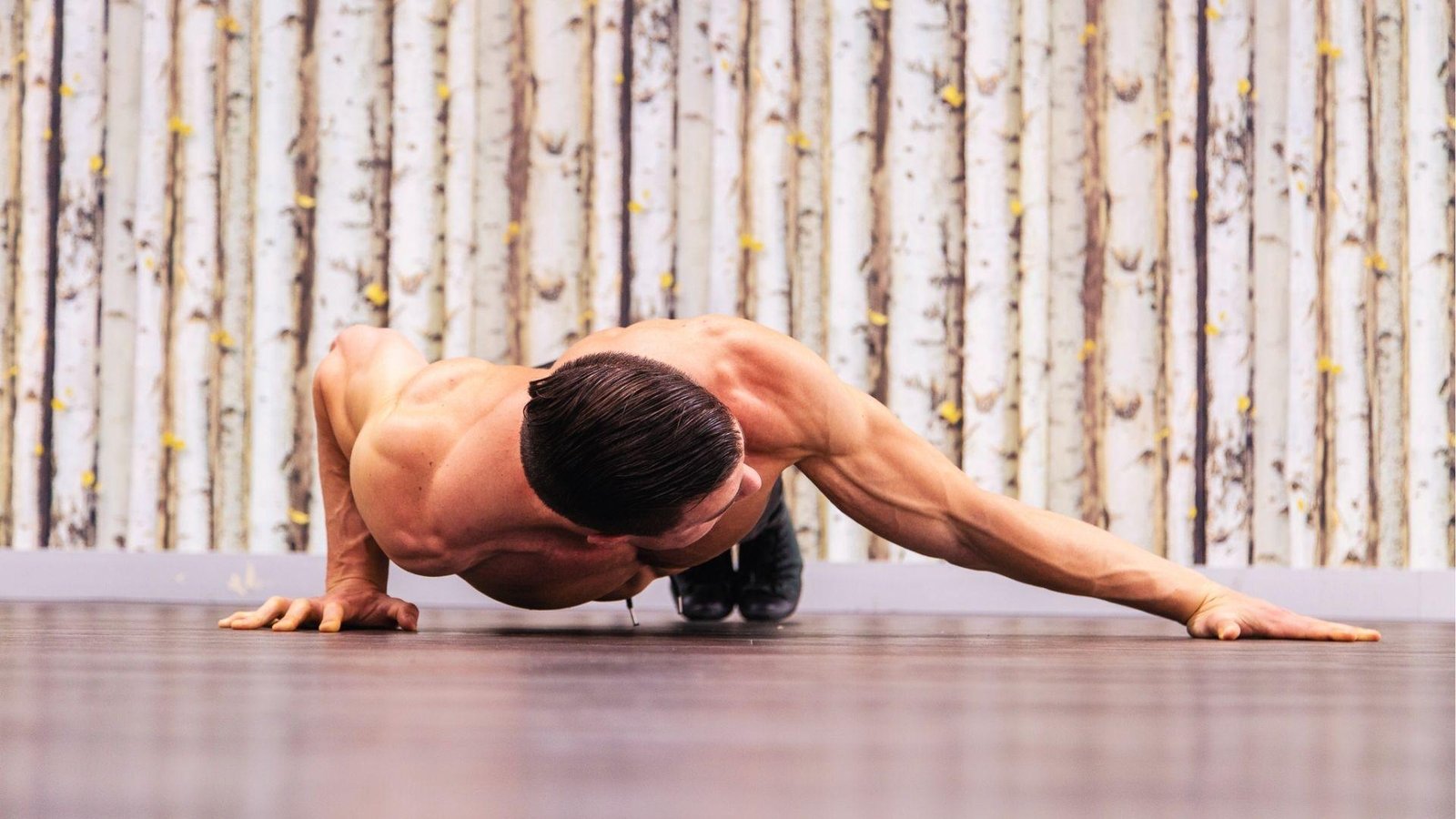 Muscular man does archer yoga push-up with one arm extended low over wooden floor indoors.