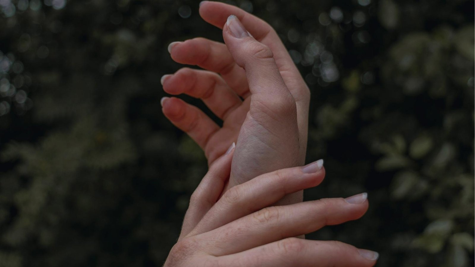 Two hands gently clasped with blurred green foliage behind, illustrating self-soothing touch and emotional grounding technique.
