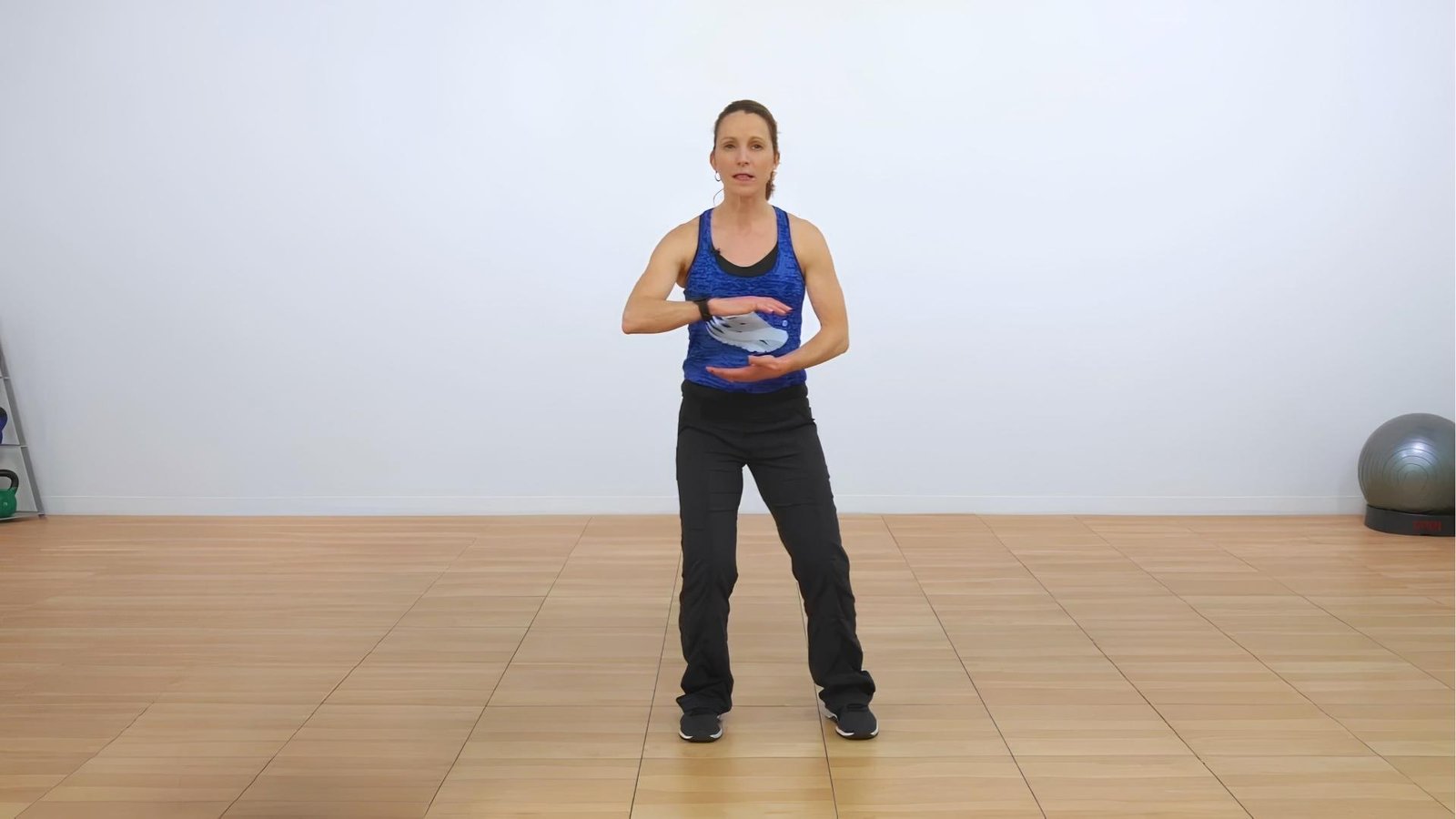 Woman in blue tank top holds "Holding the Ball" Tai Chi pose in studio, hands rounded, feet shoulder-width.