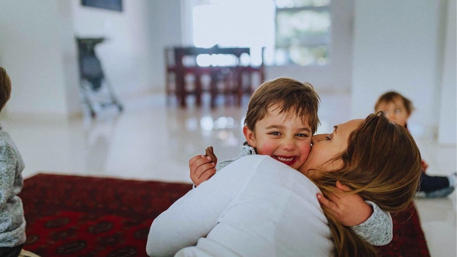 Mother in white shirt hugs smiling young boy on red rug, showing nurturing connection and emotional safety.