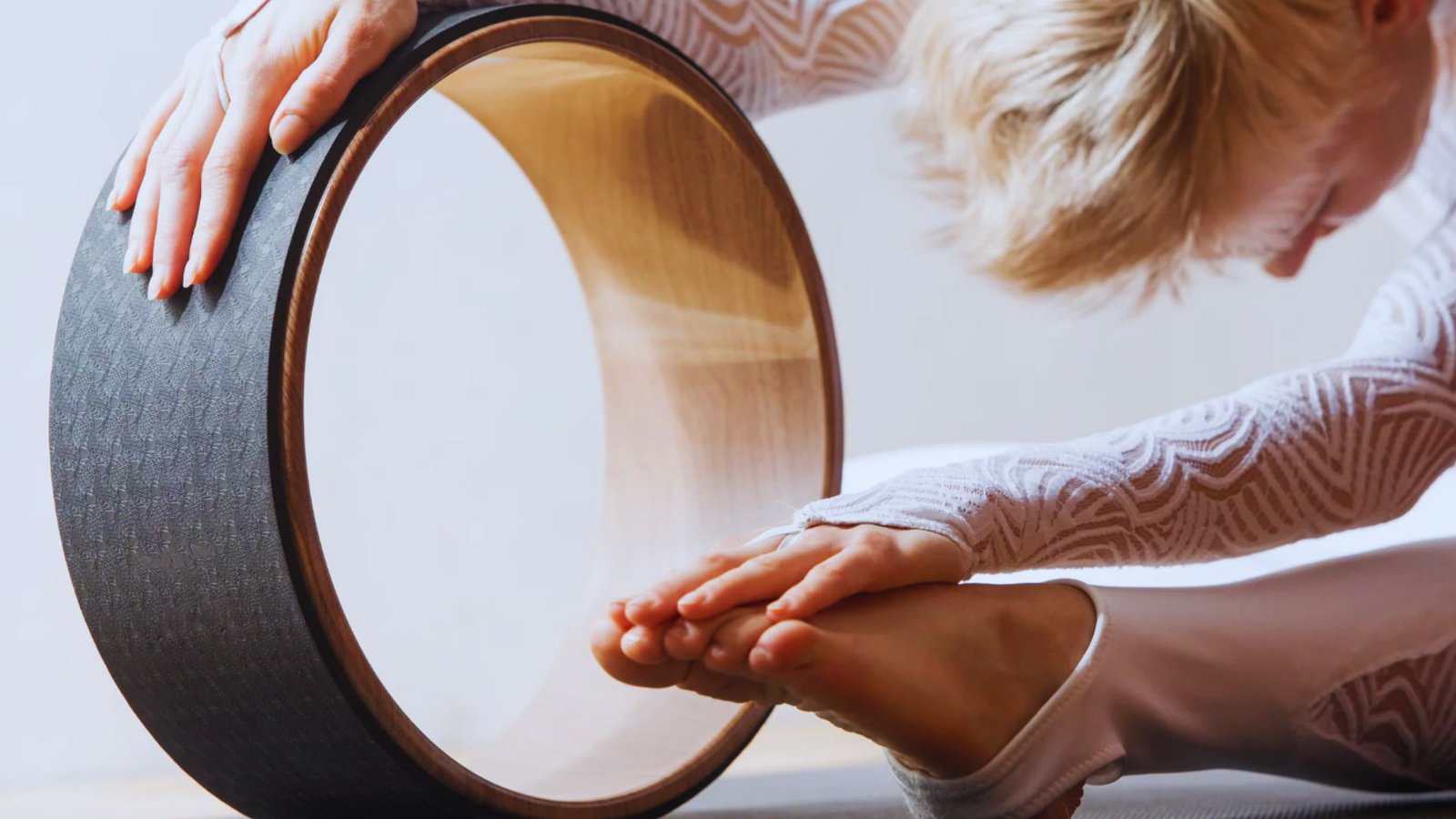 Person stretches forward while holding a yoga wheel and reaching toward feet during a seated pose.