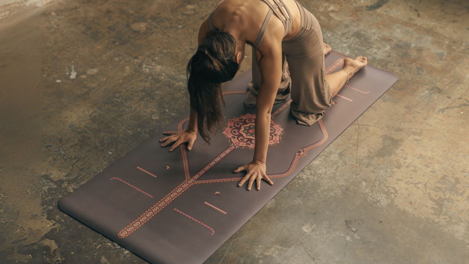 Woman in neutral yoga wear practices Cat Pose on a patterned mat with hands and knees grounded.