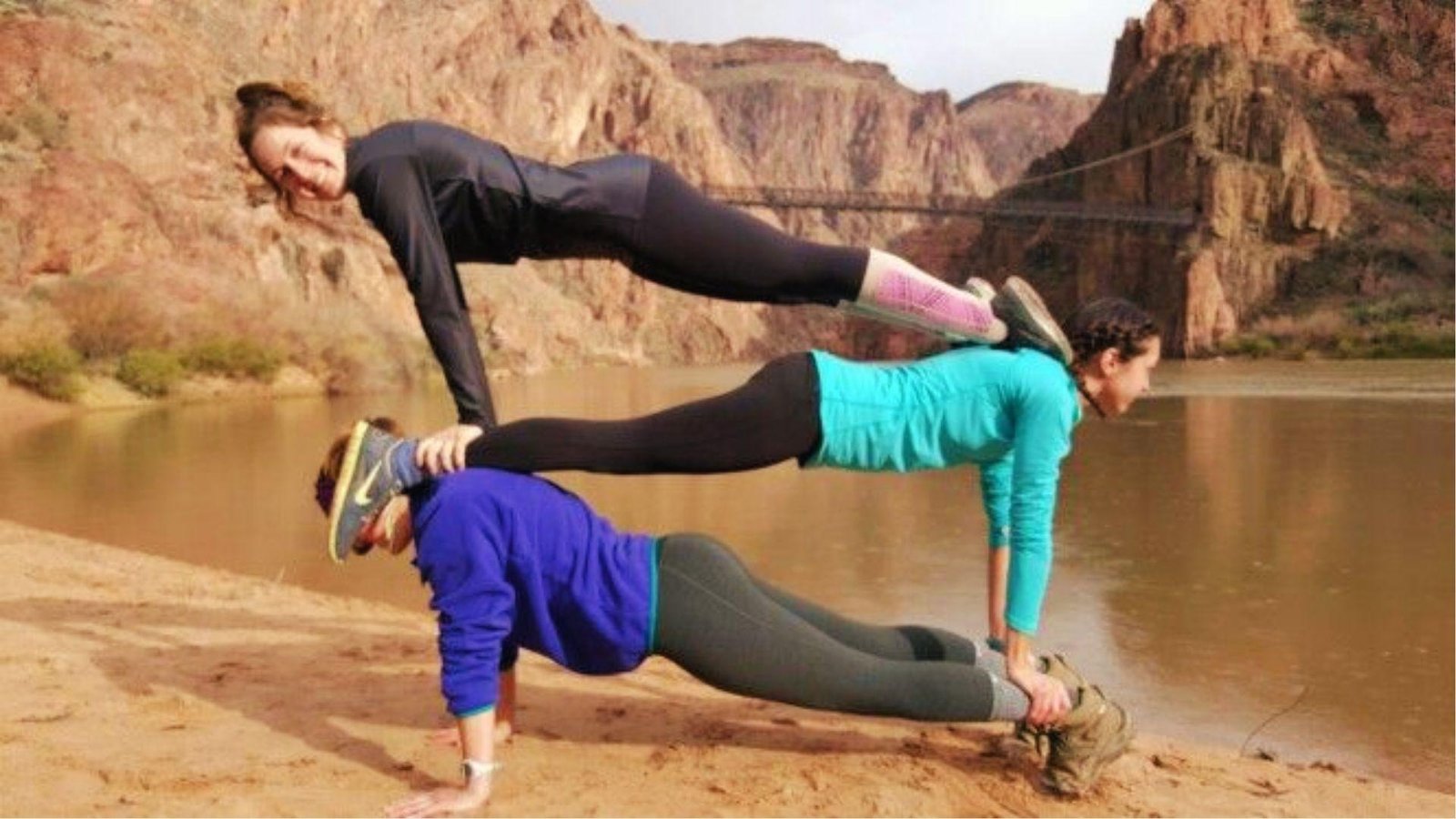 Three women stack in plank pose by a river, balancing in a 3 person yoga pyramid outdoors.