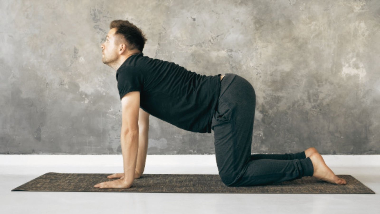 Man in black t-shirt and pants performs Cow Pose on a dark yoga mat with belly dropping and tailbone lifted.