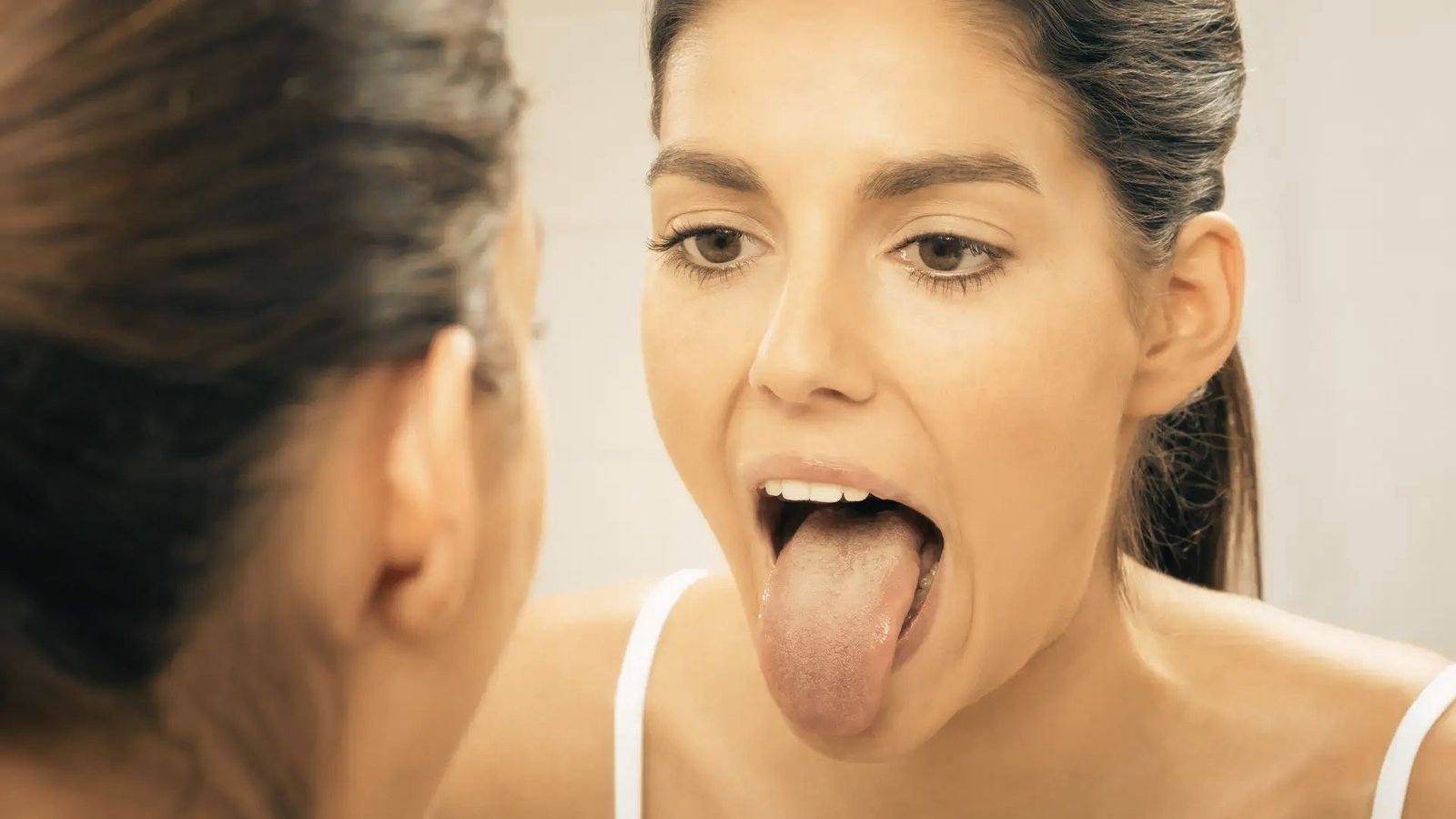 Woman looking in a mirror performs a tongue extension exercise, stretching her tongue down toward her chin.