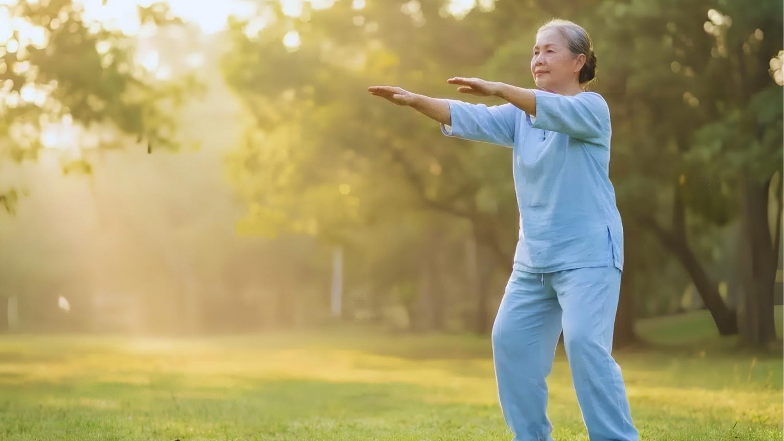 Senior woman in light blue suit practices Tai Chi in sunlit park, arms extended forward, calm balanced posture.