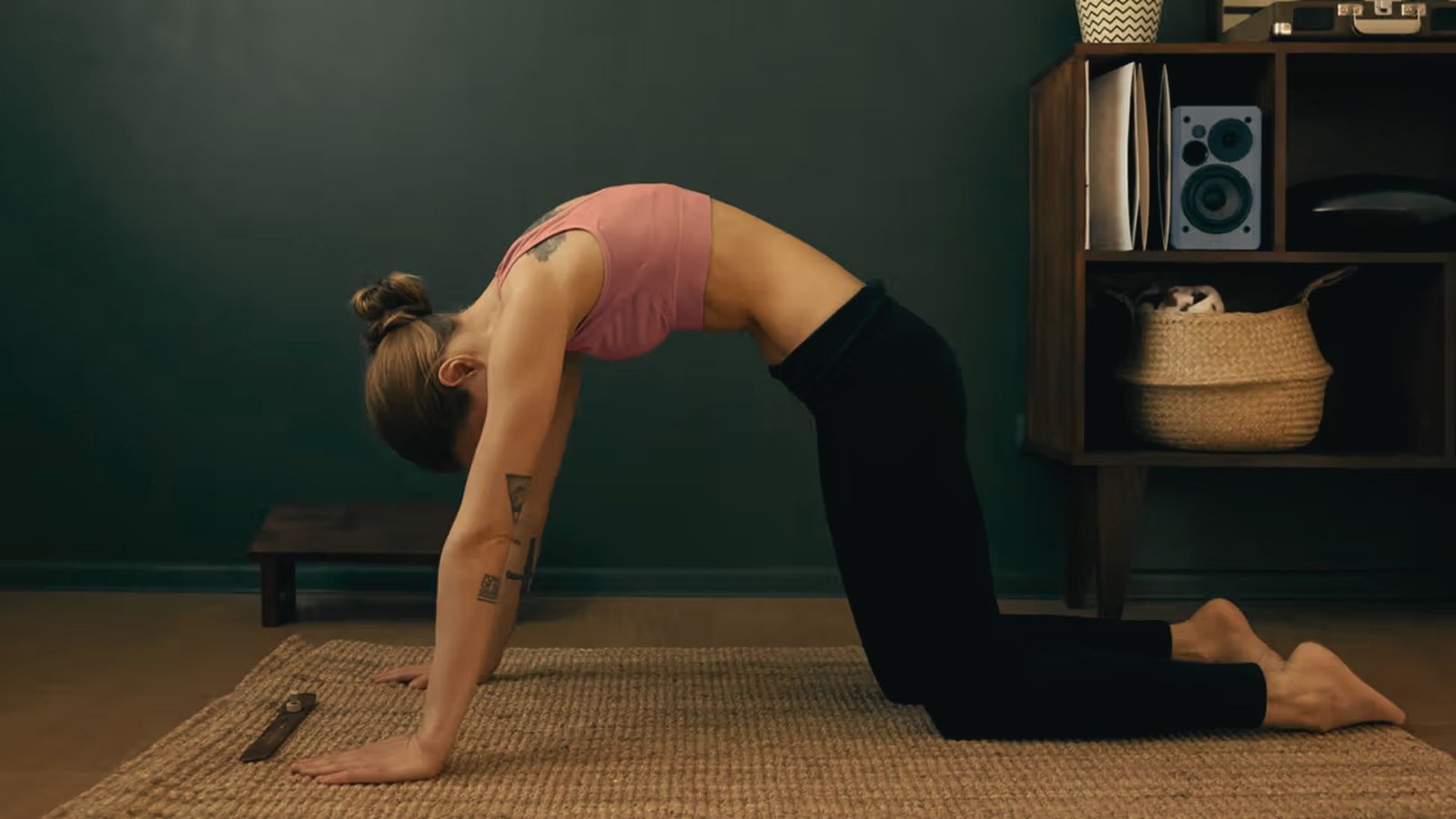 Woman in pink tank top and black leggings demonstrating Cat Pose on a woven mat indoors.
