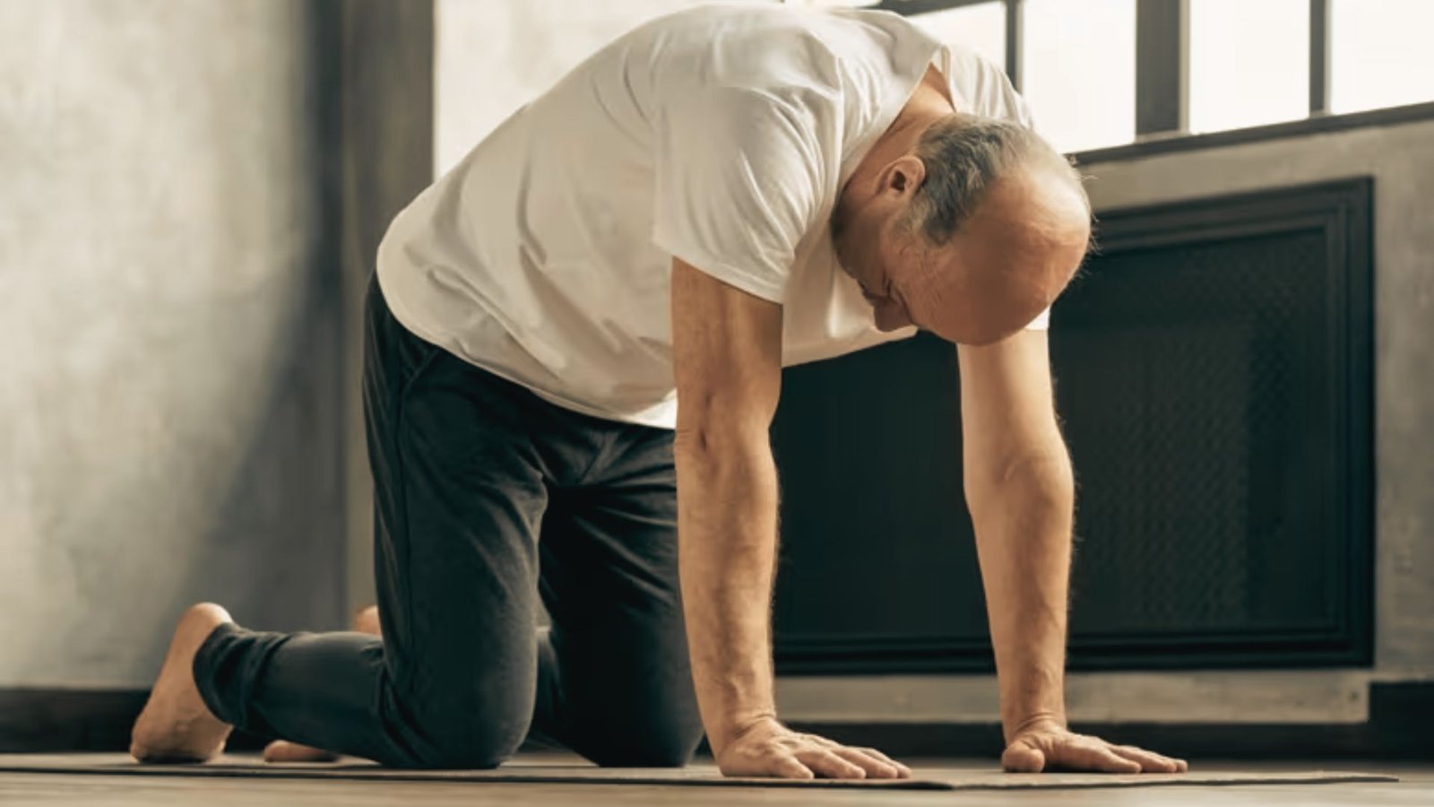 An older man in a white tee practices cat pose on a yoga mat, gently rounding his spine with focused attention.