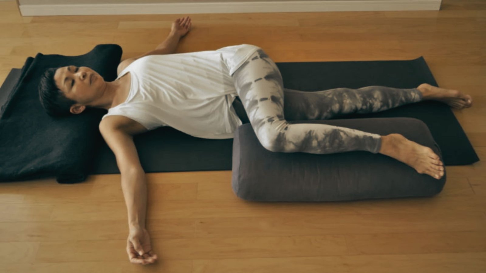 Person lying on yoga mat in supported supine twist pose, with one knee bent over a bolster and arms extended.