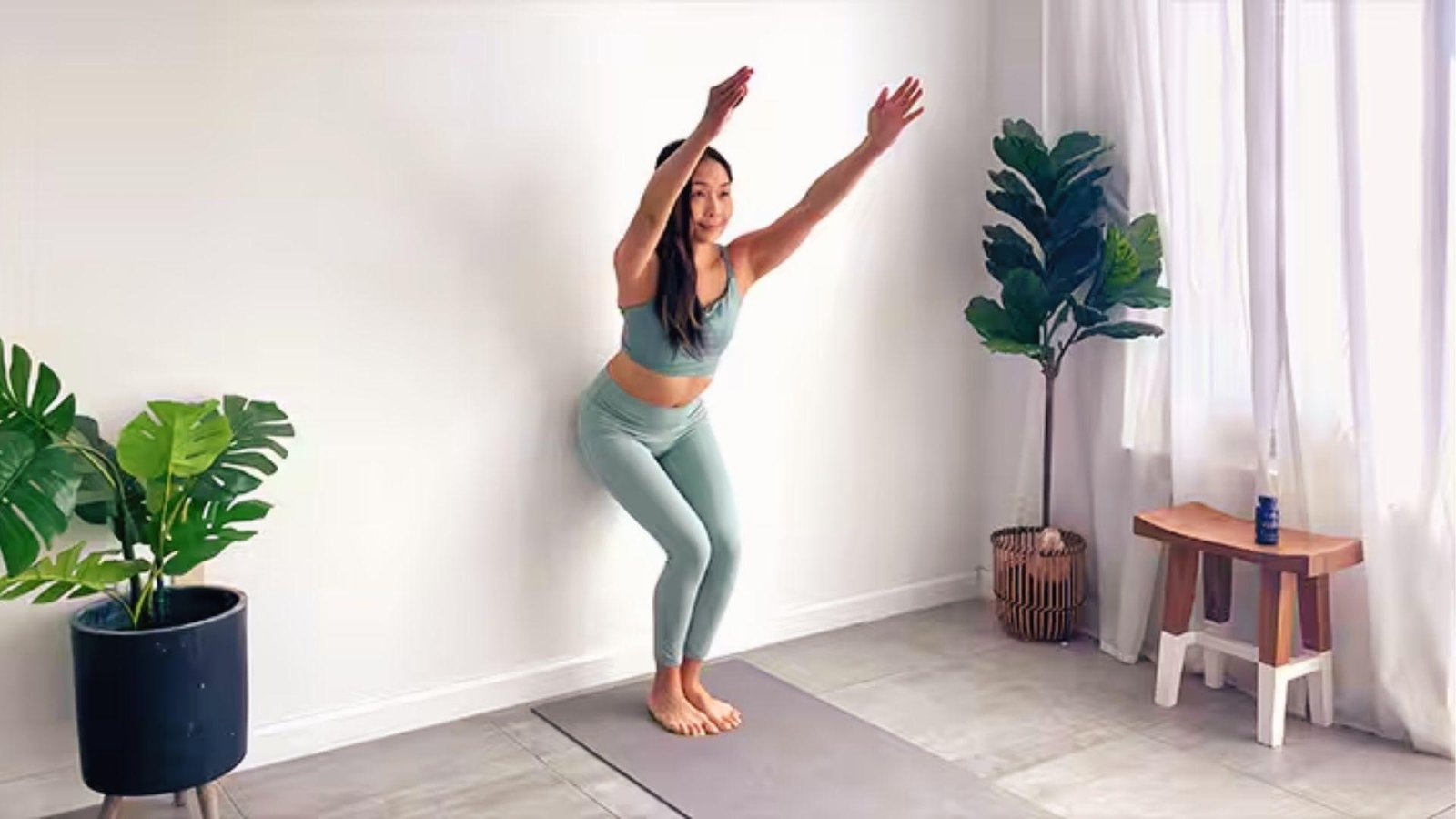 Woman in wall yoga chair pose with back on wall, arms raised, building leg and core strength.
