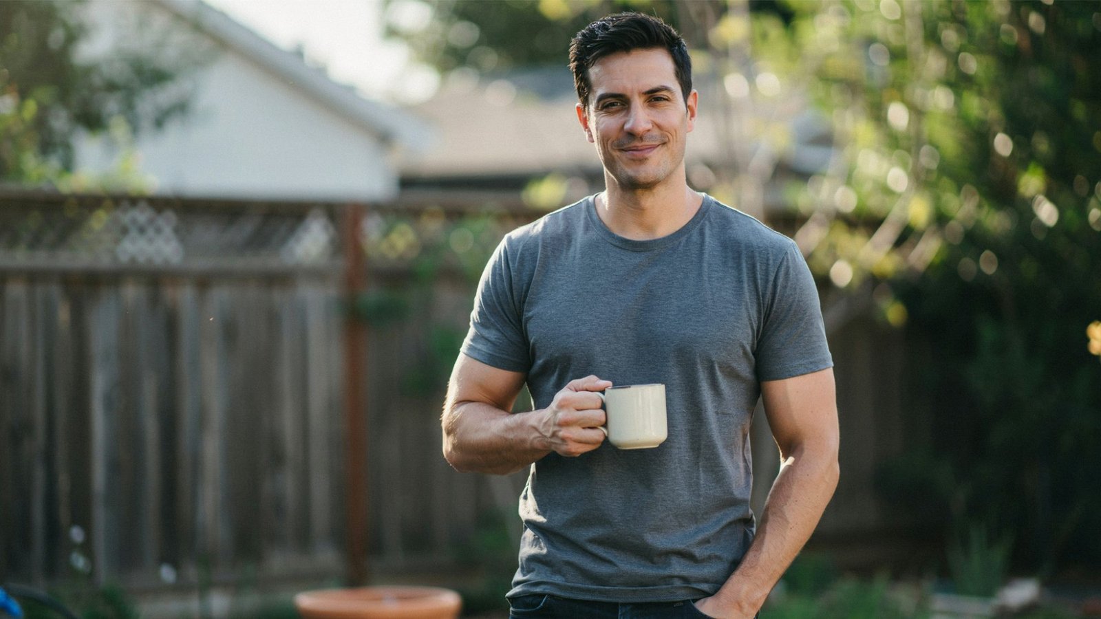 Man stands outdoors holding a mug and smiling in a backyard during soft morning sunlight.