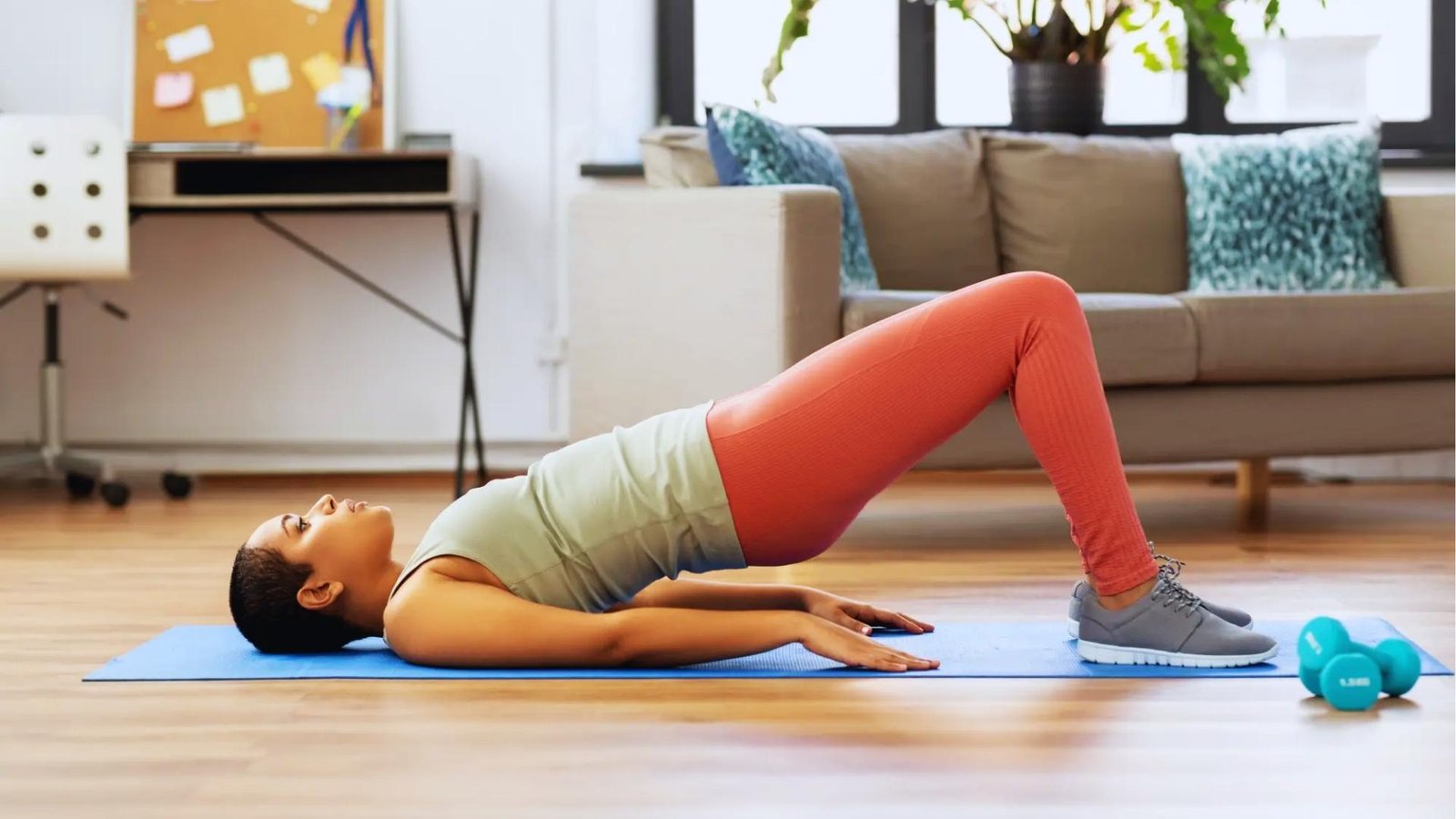 Woman in coral leggings and sage tank holds glute bridge on blue mat in living room, activating pelvic floor.