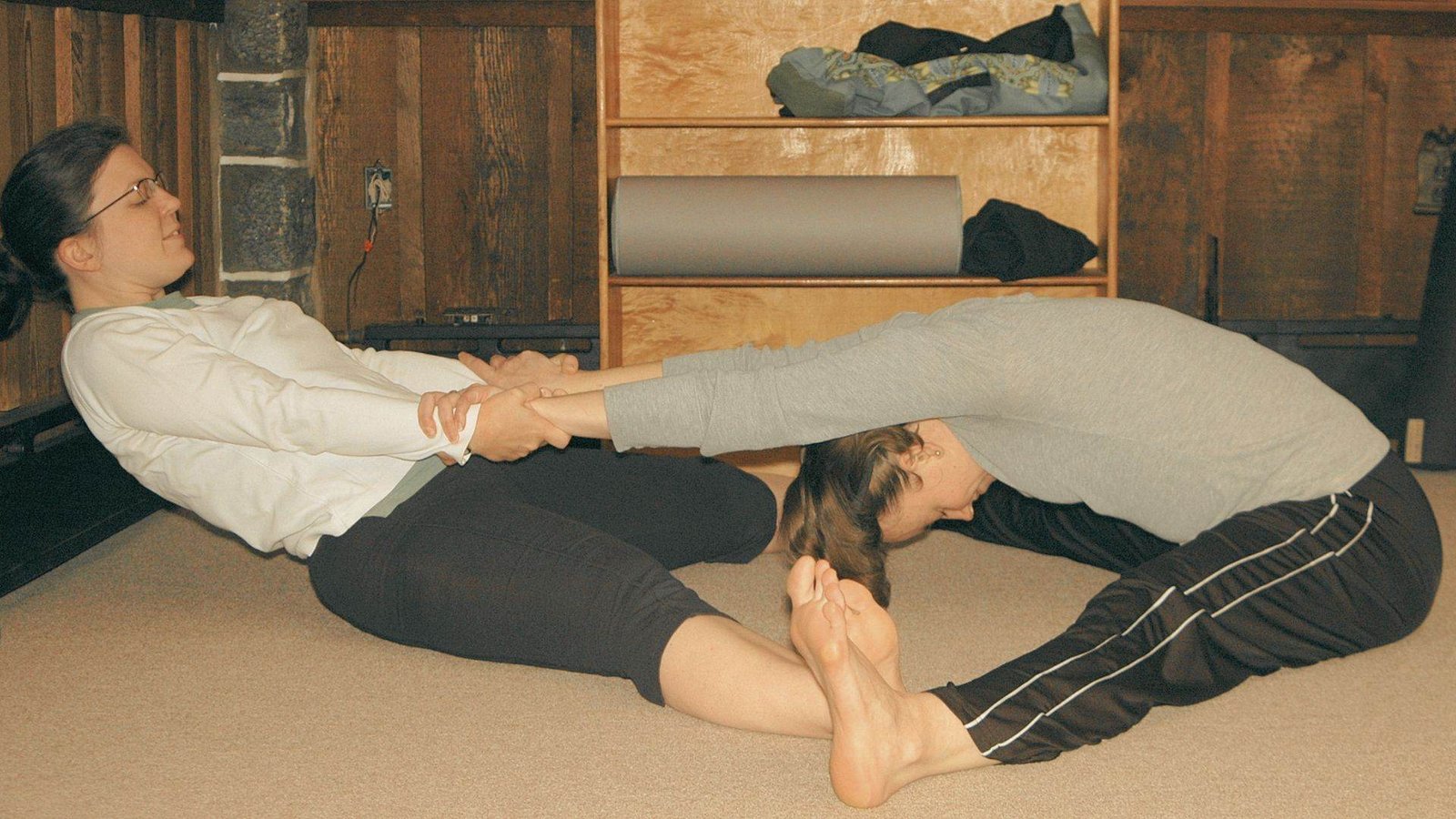 A couple sits back-to-back in a cross-legged pose on a yoga mat in a minimalist studio.
