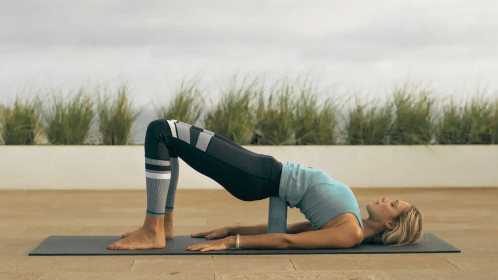 Woman lying on her back in a supported bridge pose with a yoga block under her sacrum and knees bent.