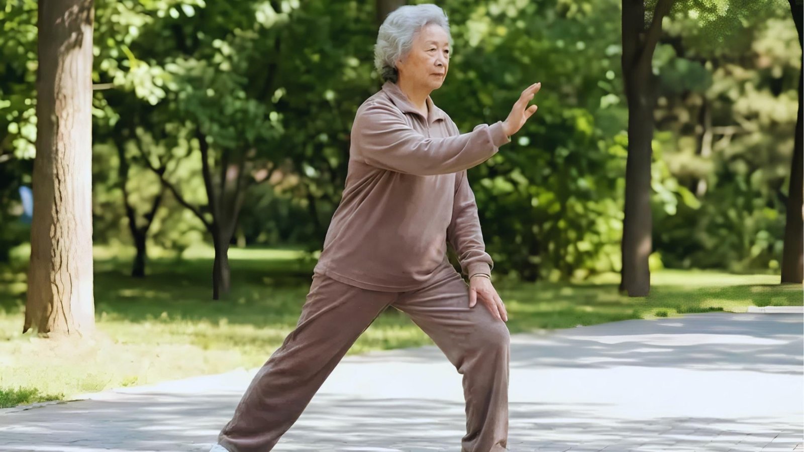 Elderly woman in taupe outfit holds low Tai Chi bow stance on park path, one hand extended, focused gaze.