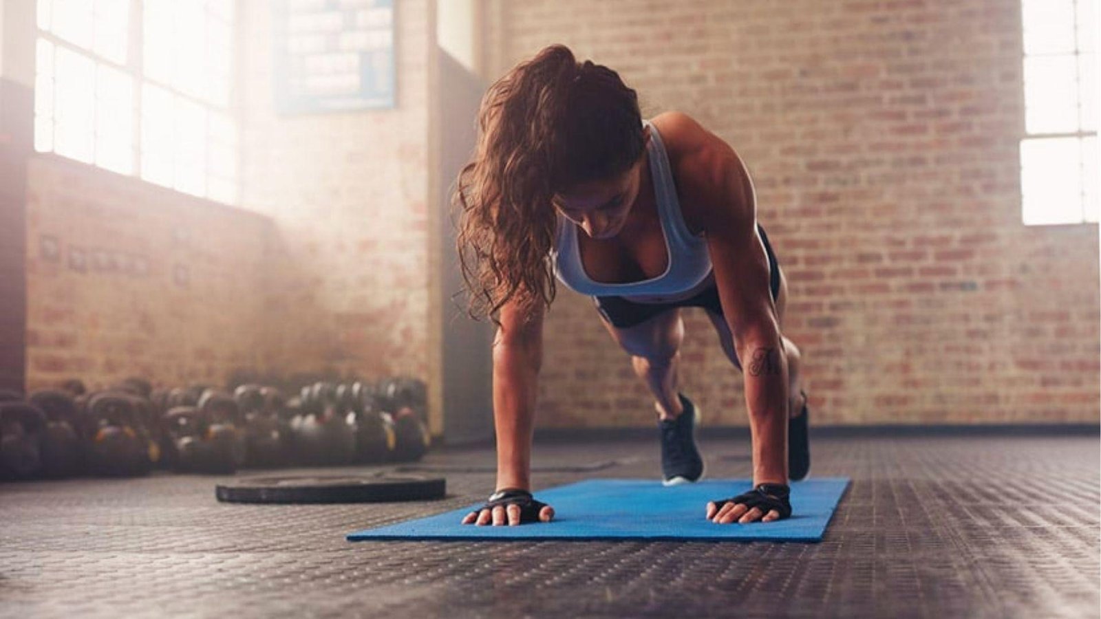An athletic woman starts yoga push-ups on a plank on a blue mat in a sunlit gym with kettlebells behind.