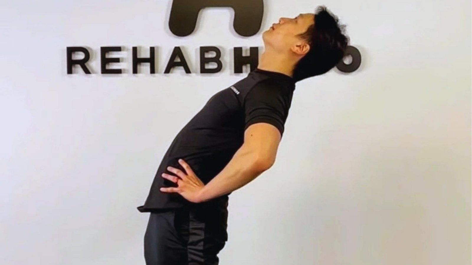 Man in black athletic wear performs standing backbend with hands on hips, arching spine to stretch in rehab studio.