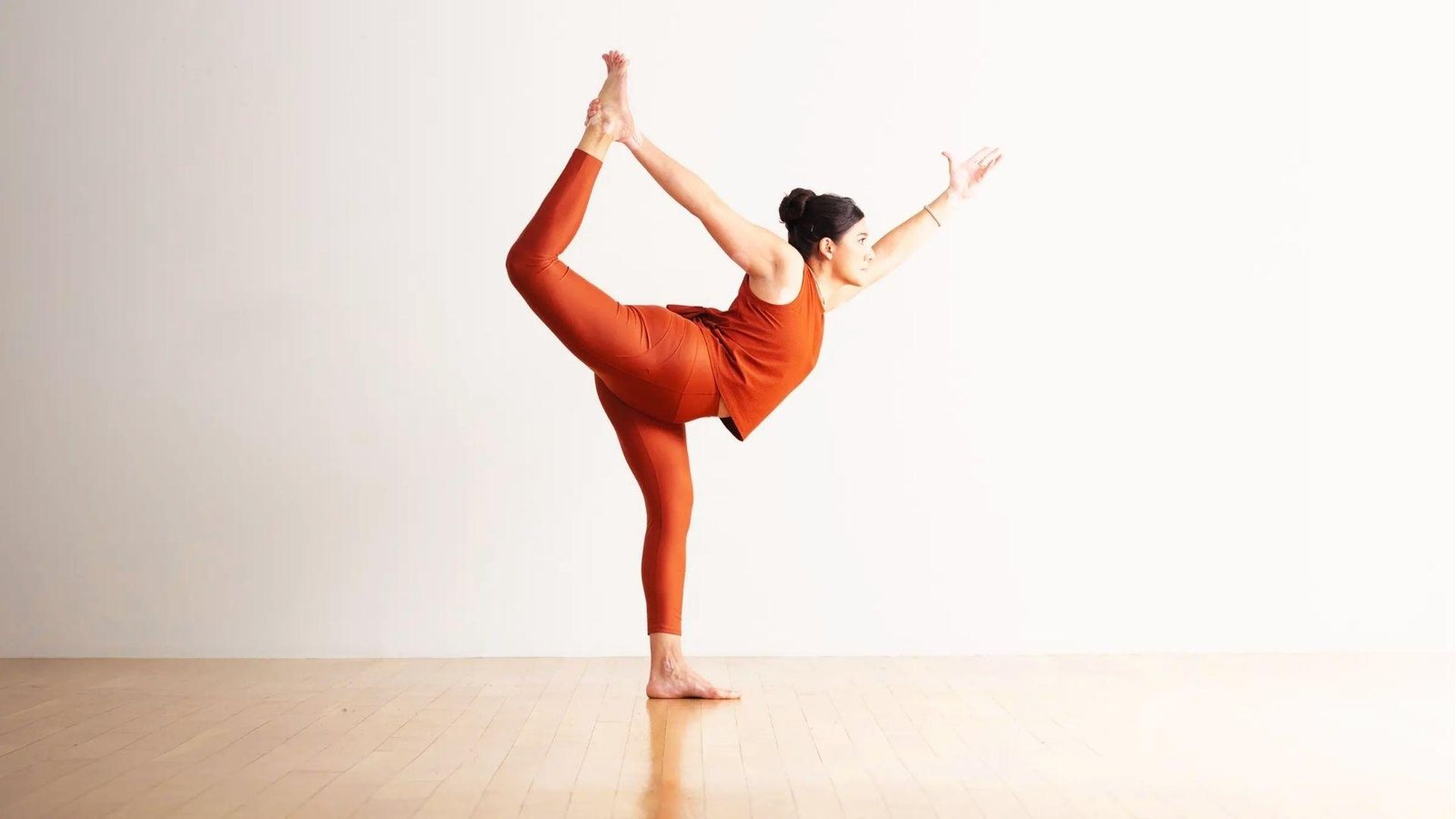 A woman in rust set dancer poses in a bright studio, white wall and wood floor visible.