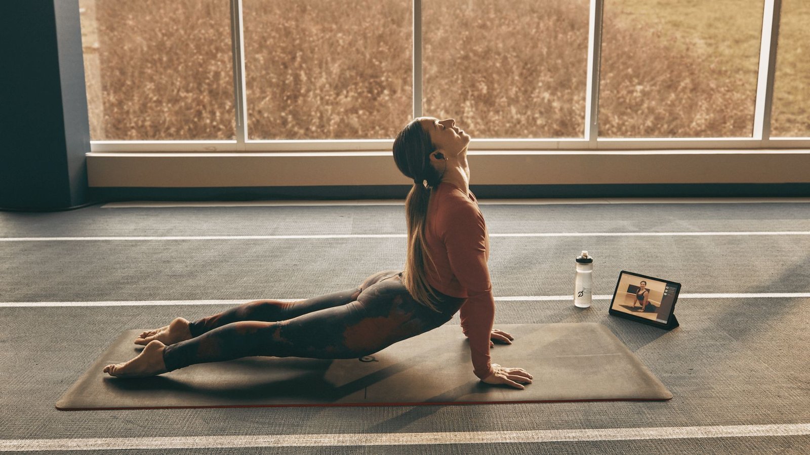 A woman in a rust-colored top lifts into cobra pose on a mat, following an online class near large windows.
