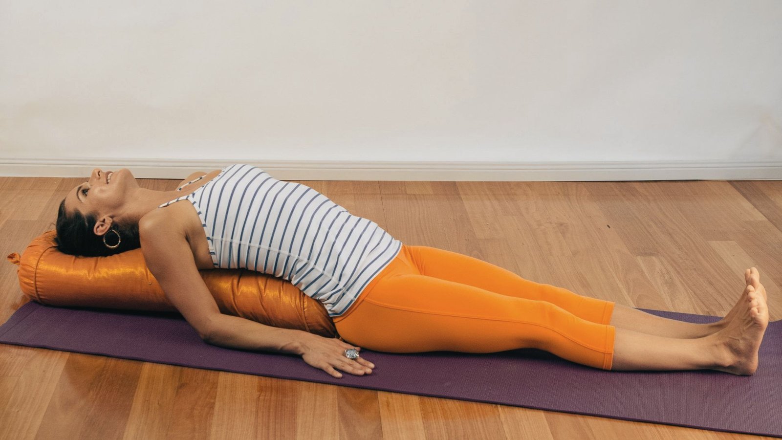 A woman lies on a purple mat with an orange bolster supporting her spine in a gentle heart-opening backbend.