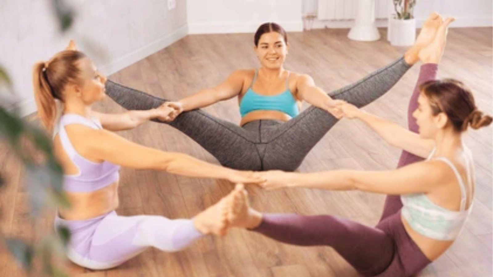 Three women sit in a circle on the floor, holding hands with legs out for 3 person yoga stretches.