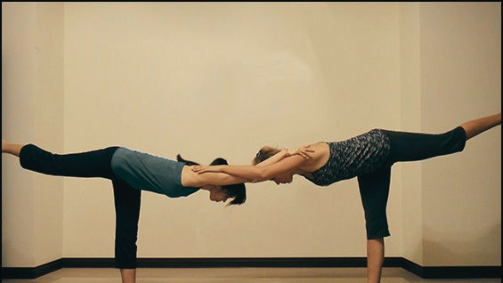 Two women balance on one leg in a yoga studio, hinging forward with arms extended to hold each other's shoulders.