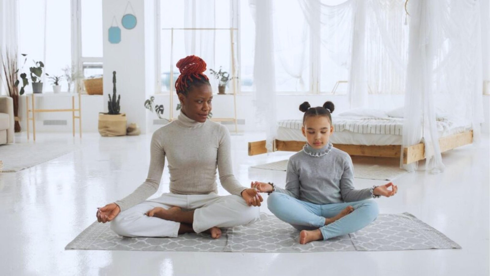 Woman with red braided bun meditates with young girl in bright white room, modeling mindfulness together.