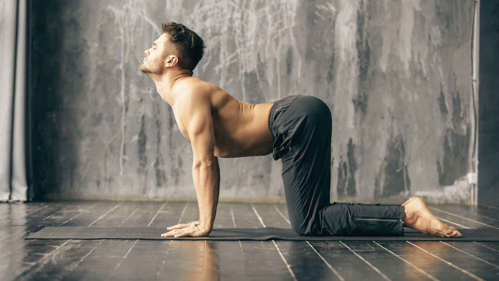 Man shirtless in black pants holds Cow Pose (Bitilasana) on a yoga mat with back arched and chest lifted.