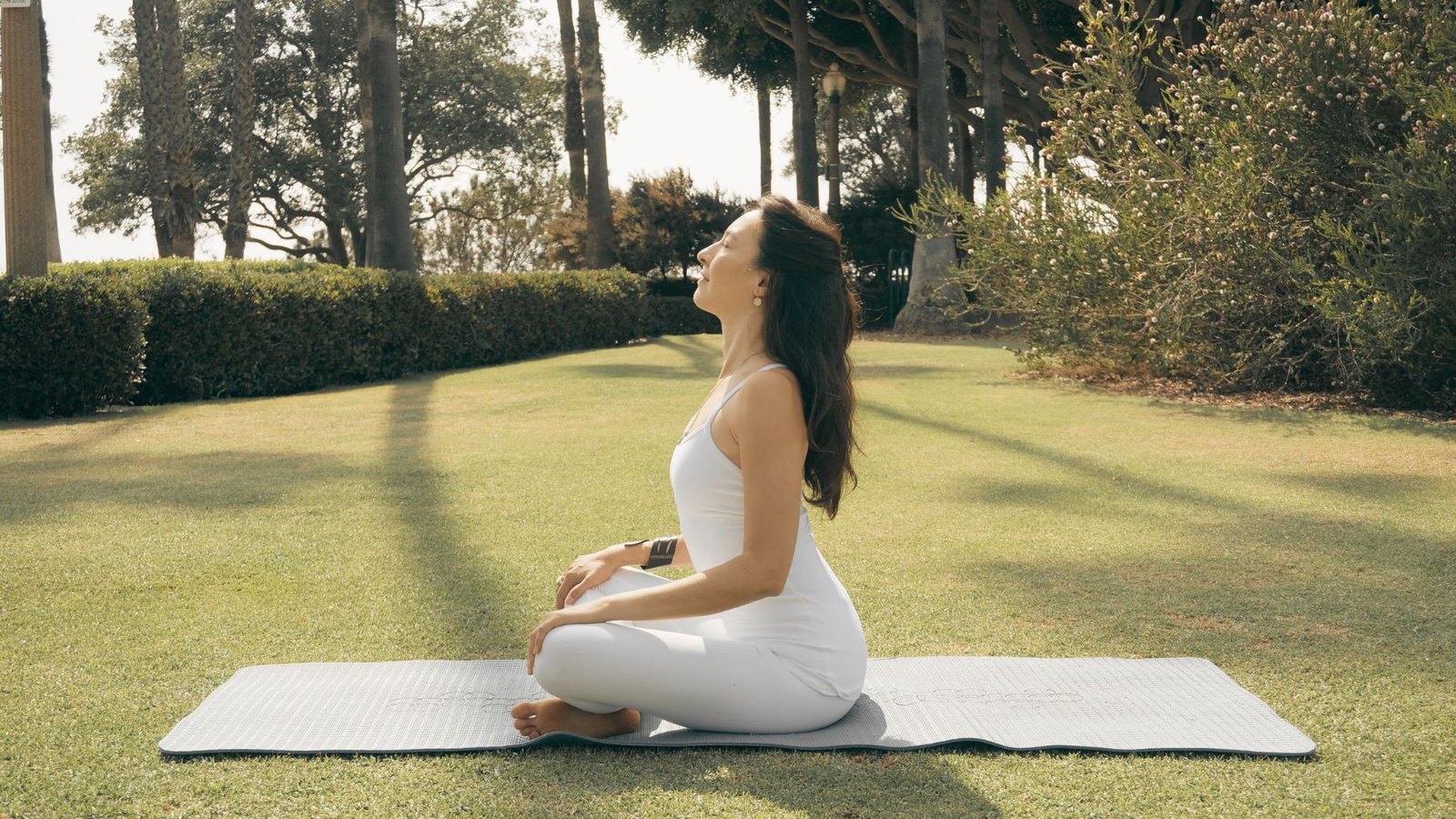 A woman in white activewear sits cross-legged on a yoga mat in a sunny park, eyes closed, head tilted back.