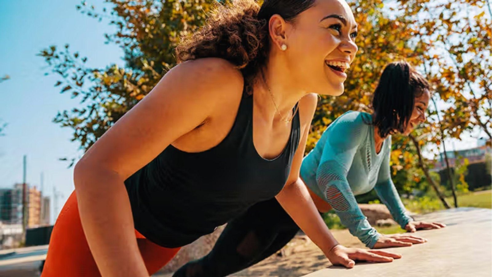 Two smiling women do incline yoga push-ups on a park bench outdoors under sunny autumn trees.