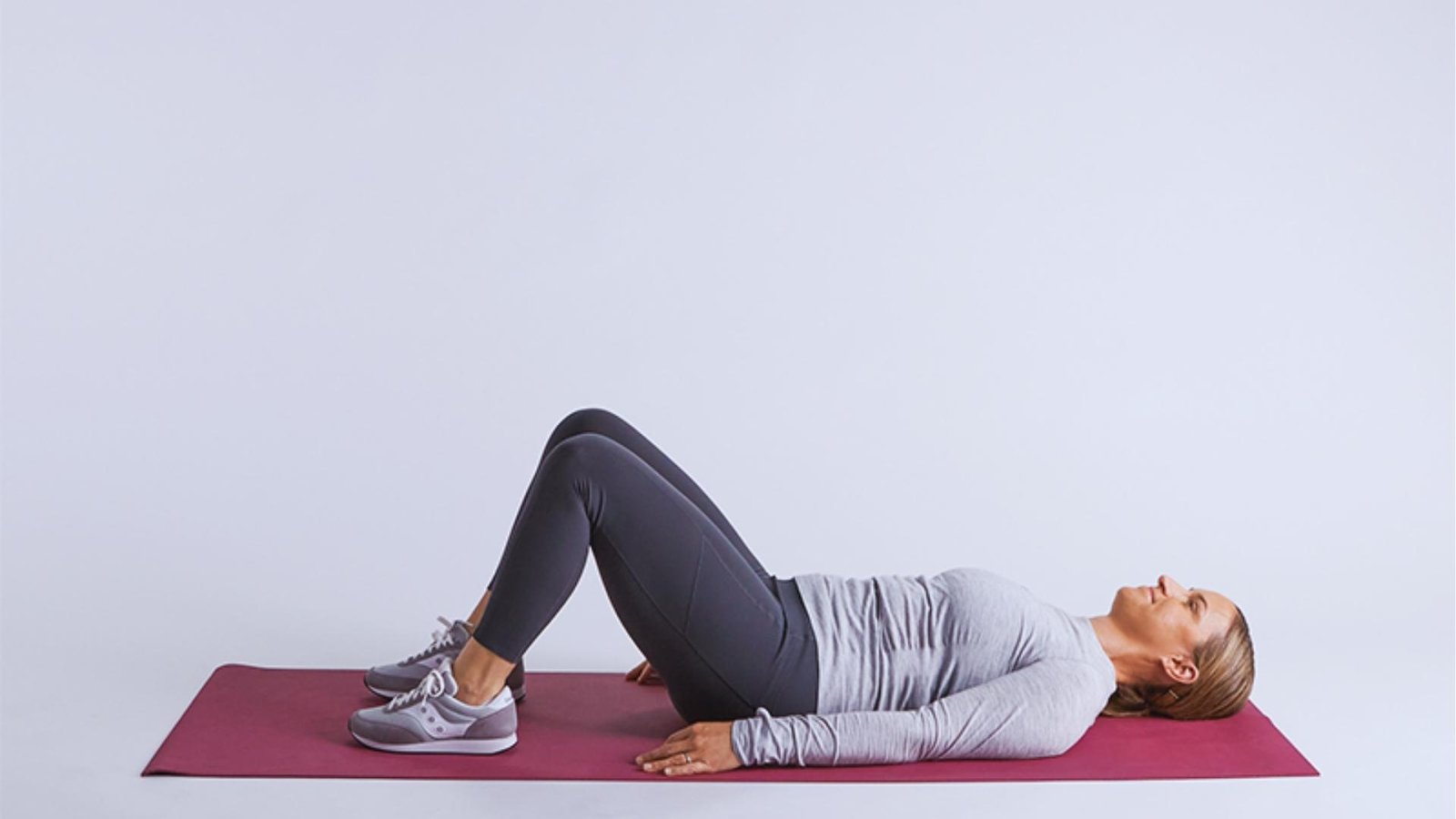 Woman in gray long-sleeve lies on mat with knees bent, preparing for pelvic tilt to engage pelvic floor.
