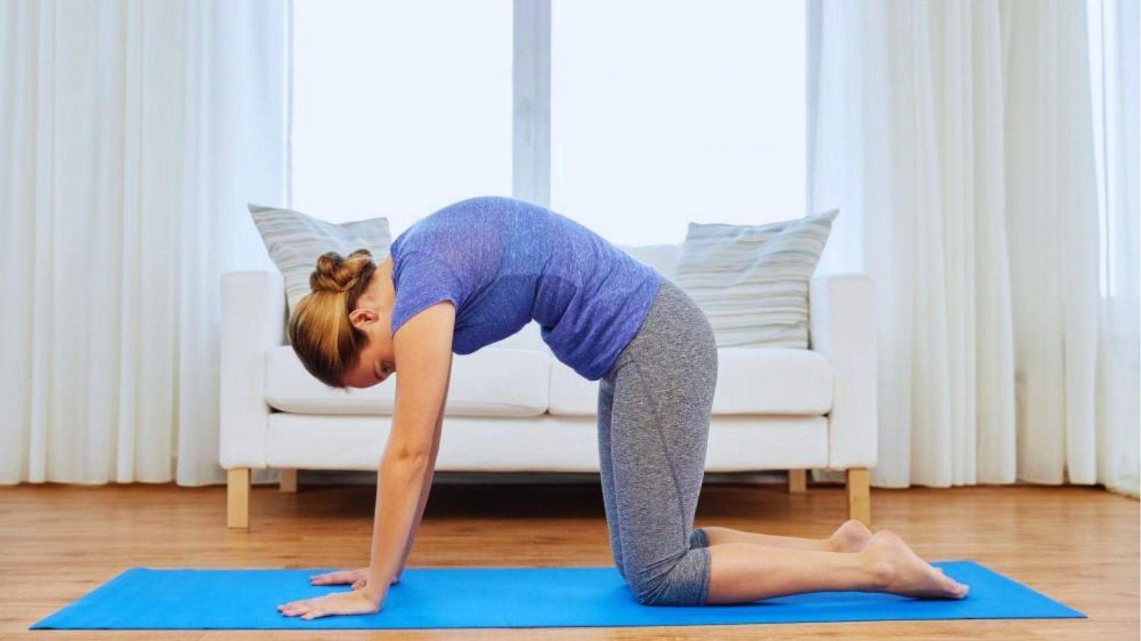 Woman in blue shirt demonstrates Cat Pose with rounded spine on blue mat, hands and knees grounded for flexion.