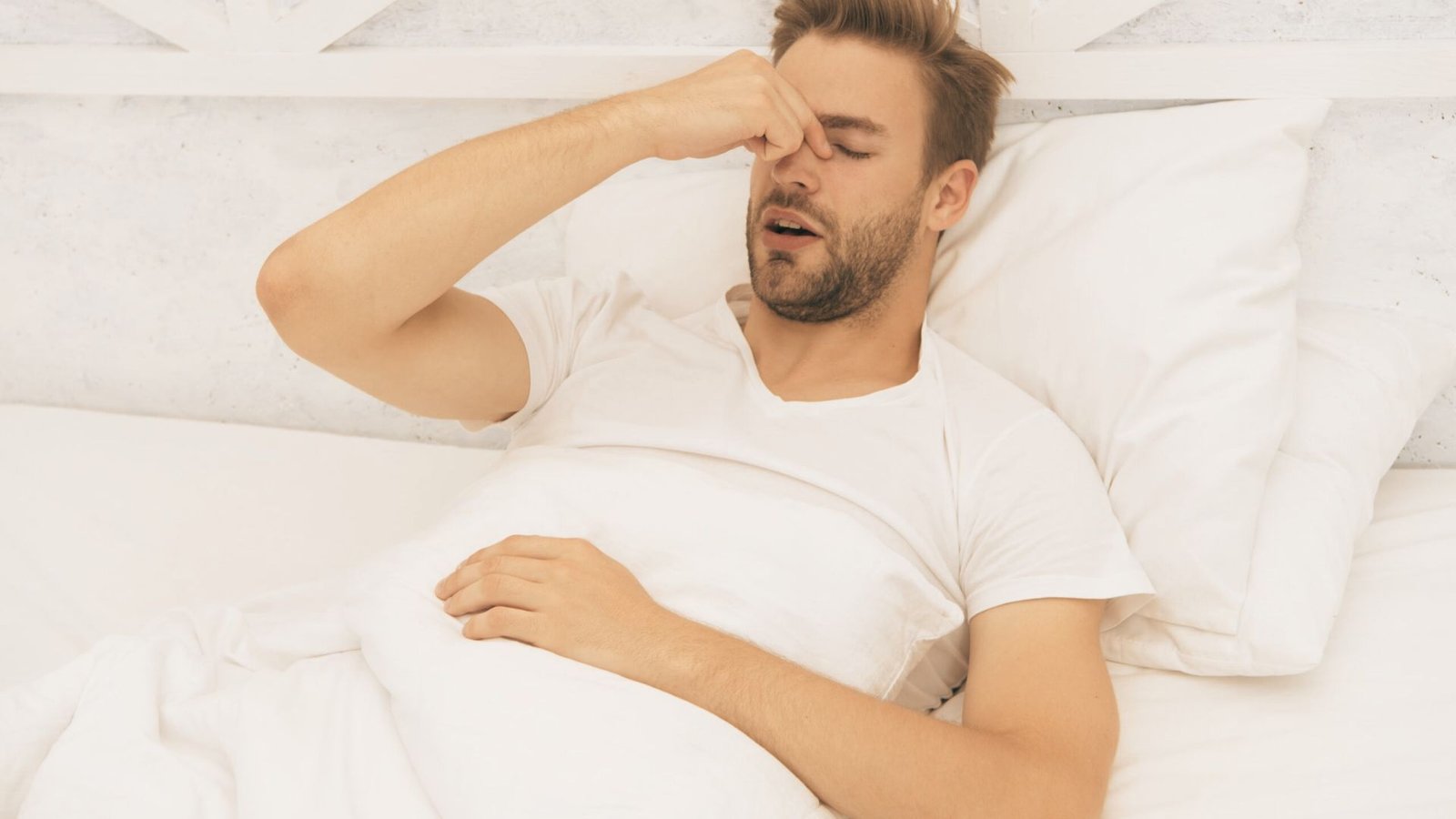 Man lying in bed with eyes closed, hand on nose, appearing fatigued from poor sleep.