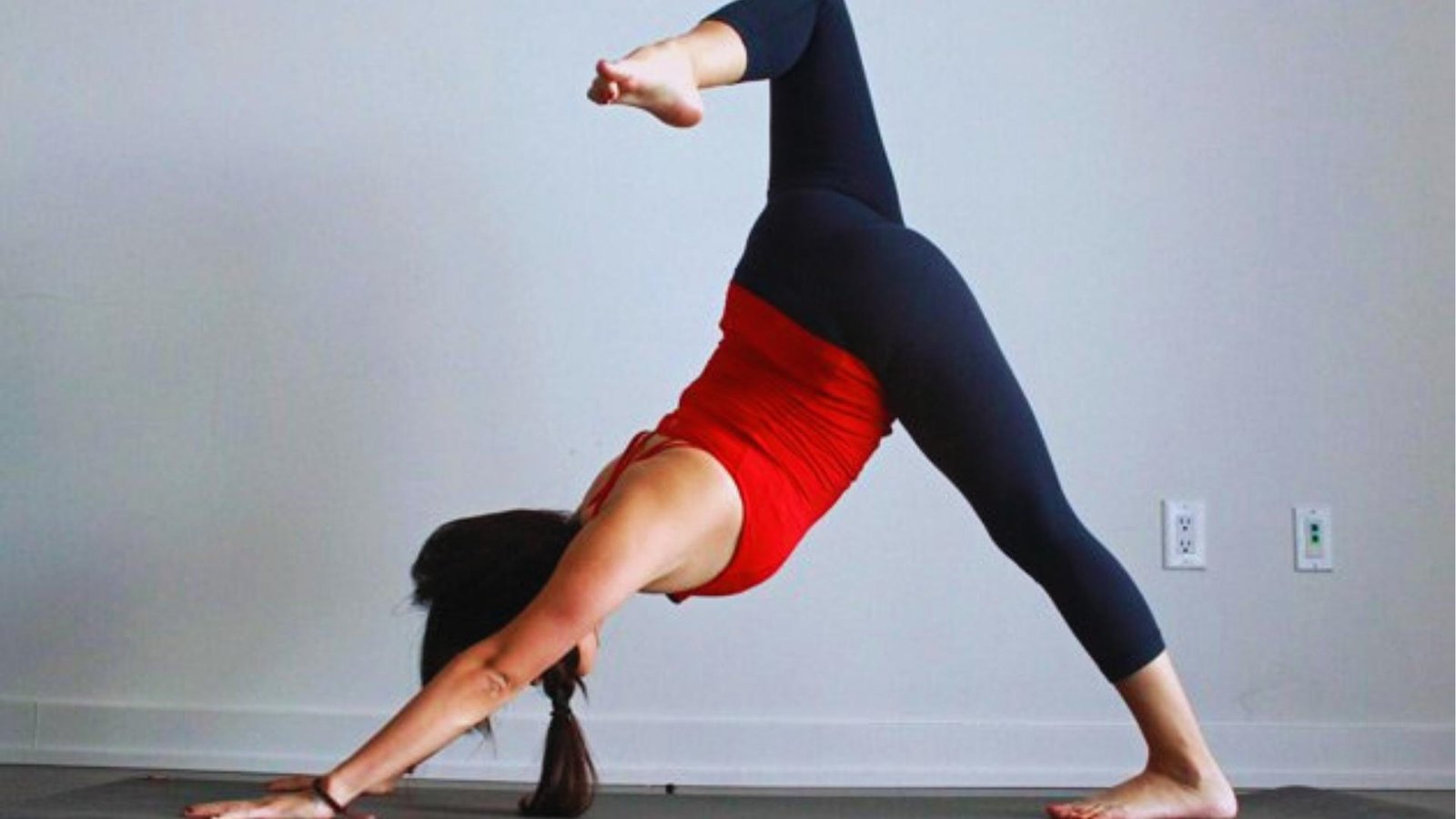 Woman in red top performs Three-Legged Downward Dog on mat, lifting leg high to open hips and lengthen spine.