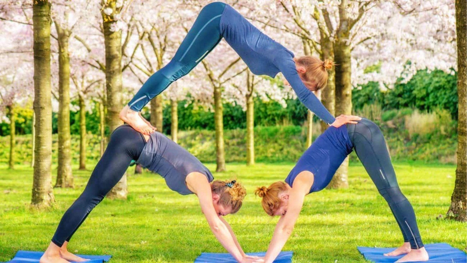 Three women form a triangle with downward dog bases, top in bridge for 3 person yoga outdoors.