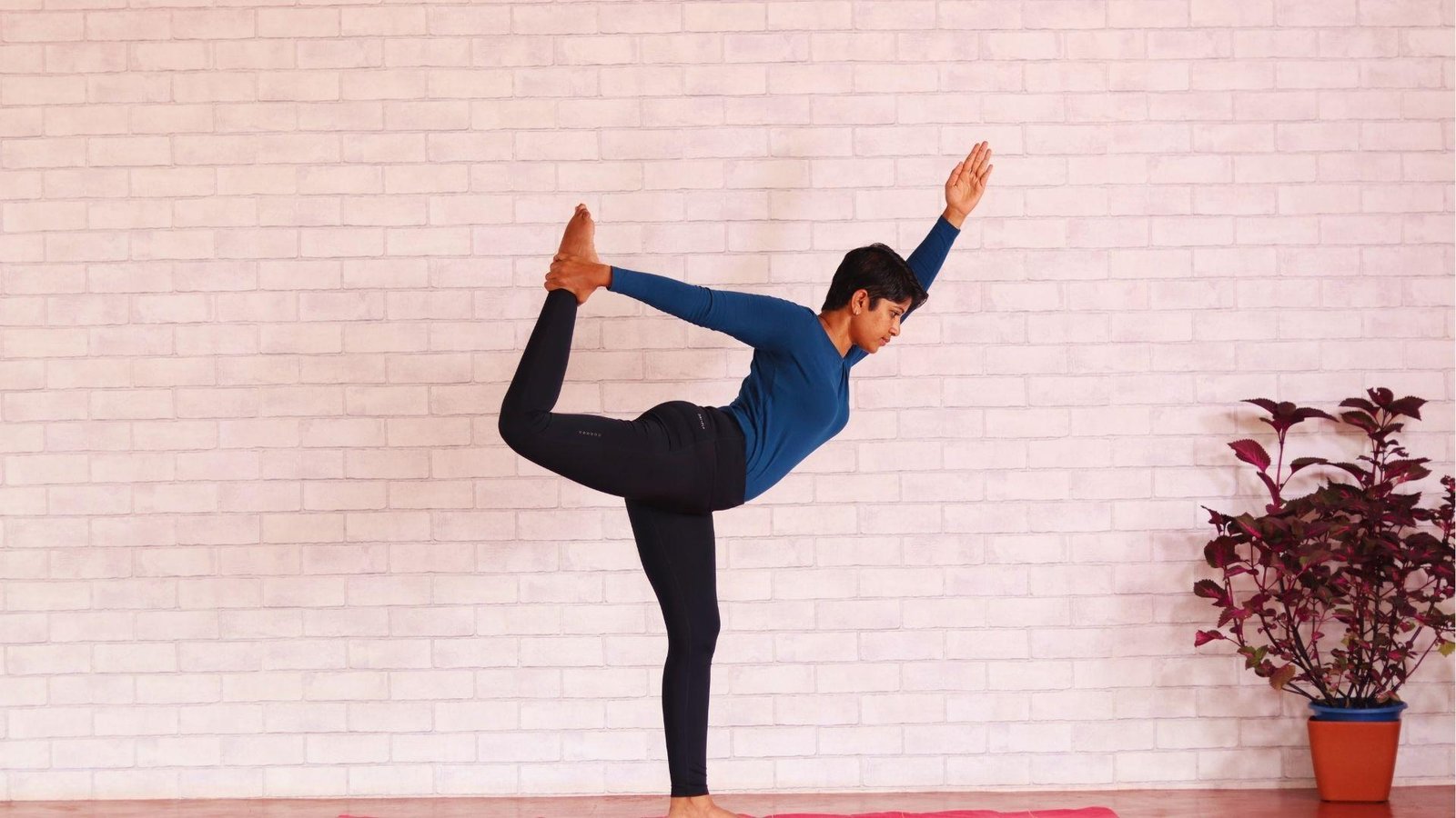 A woman in blue top holds Natarajasana indoors against white brick wall, plant to side.