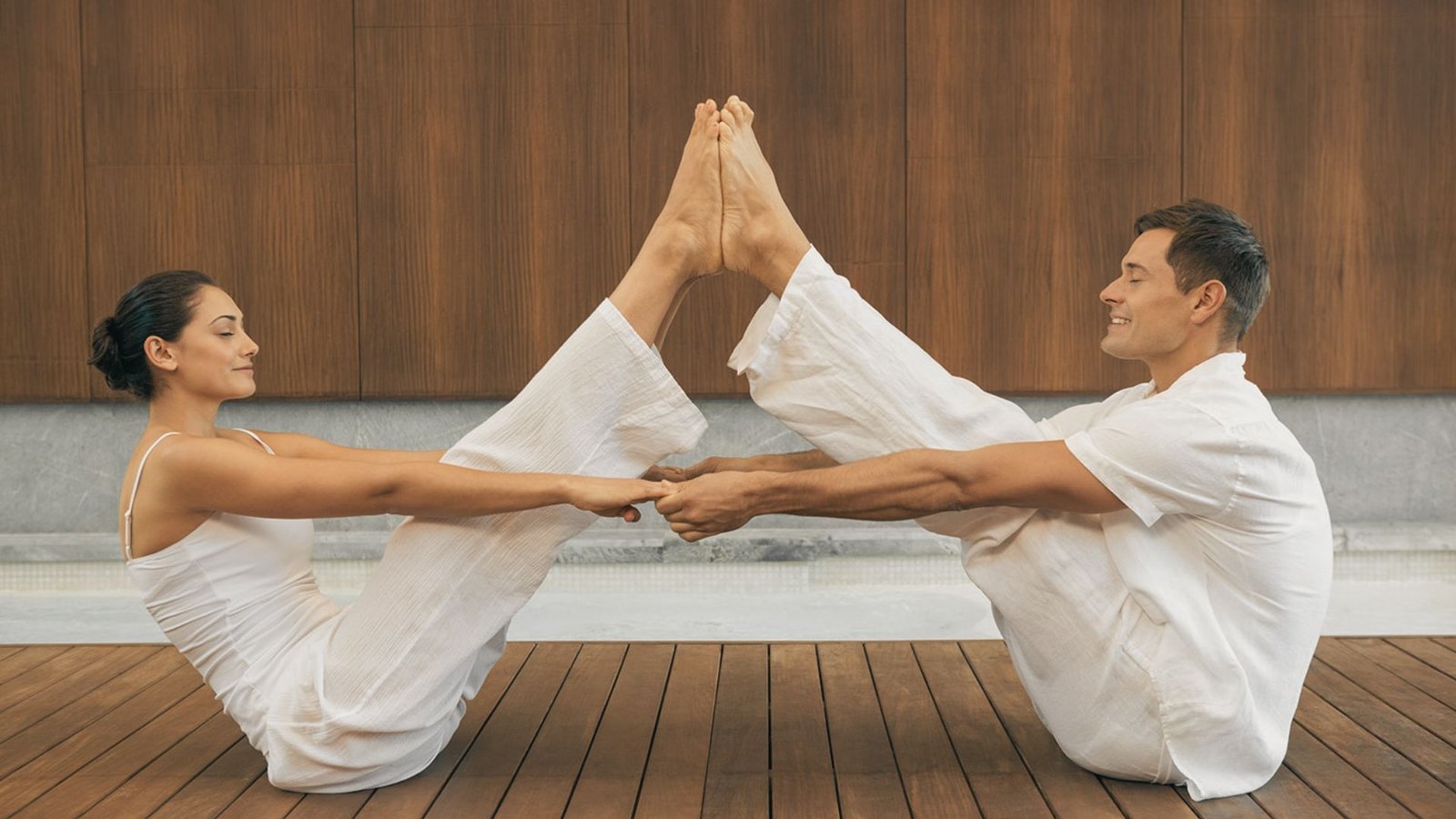 A man and woman sit facing each other on a wooden deck, holding hands with legs extended and soles touching.