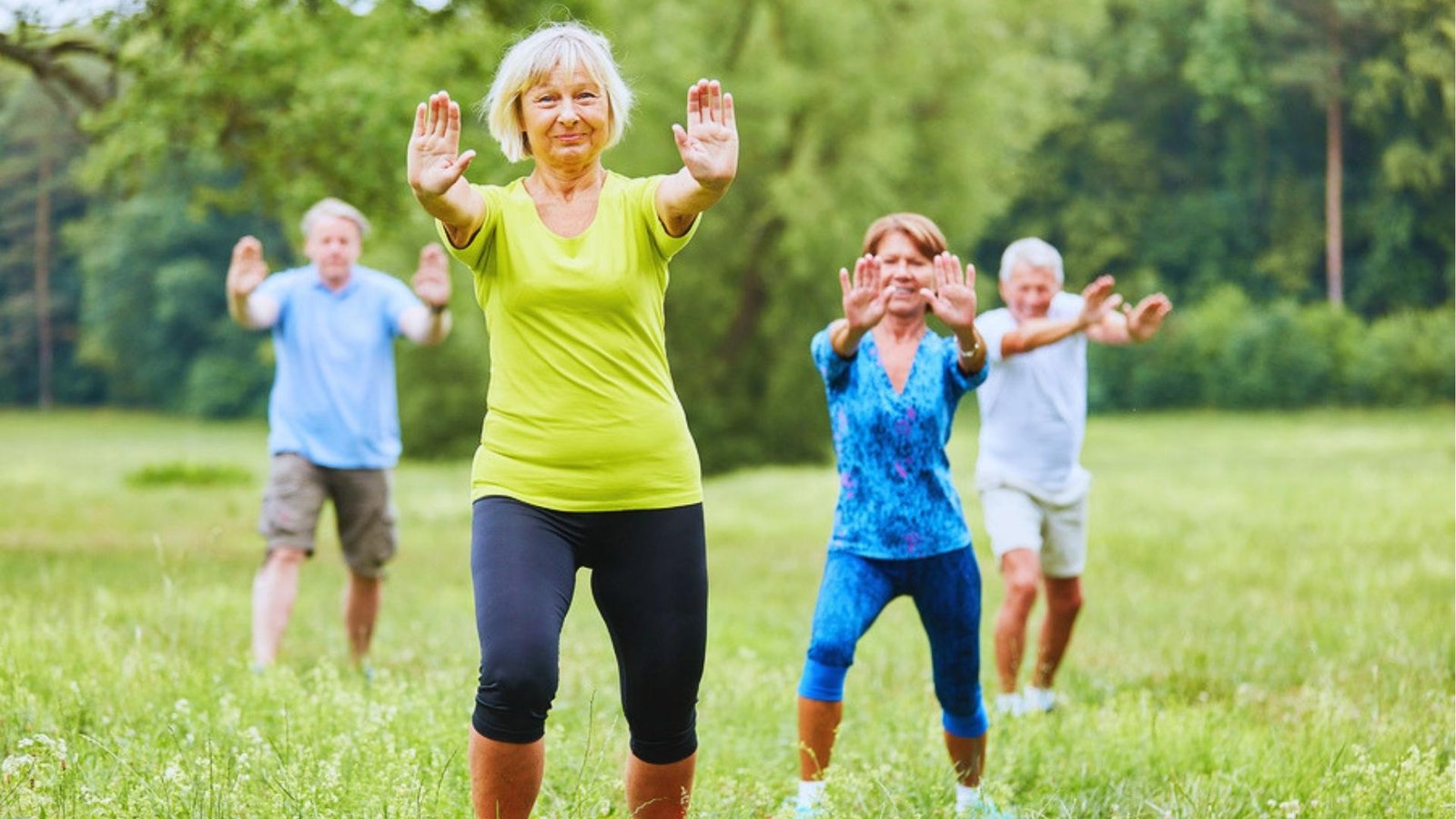 Group of four seniors practice Tai Chi in park, woman in lime shirt leads with palms forward in balanced stance.