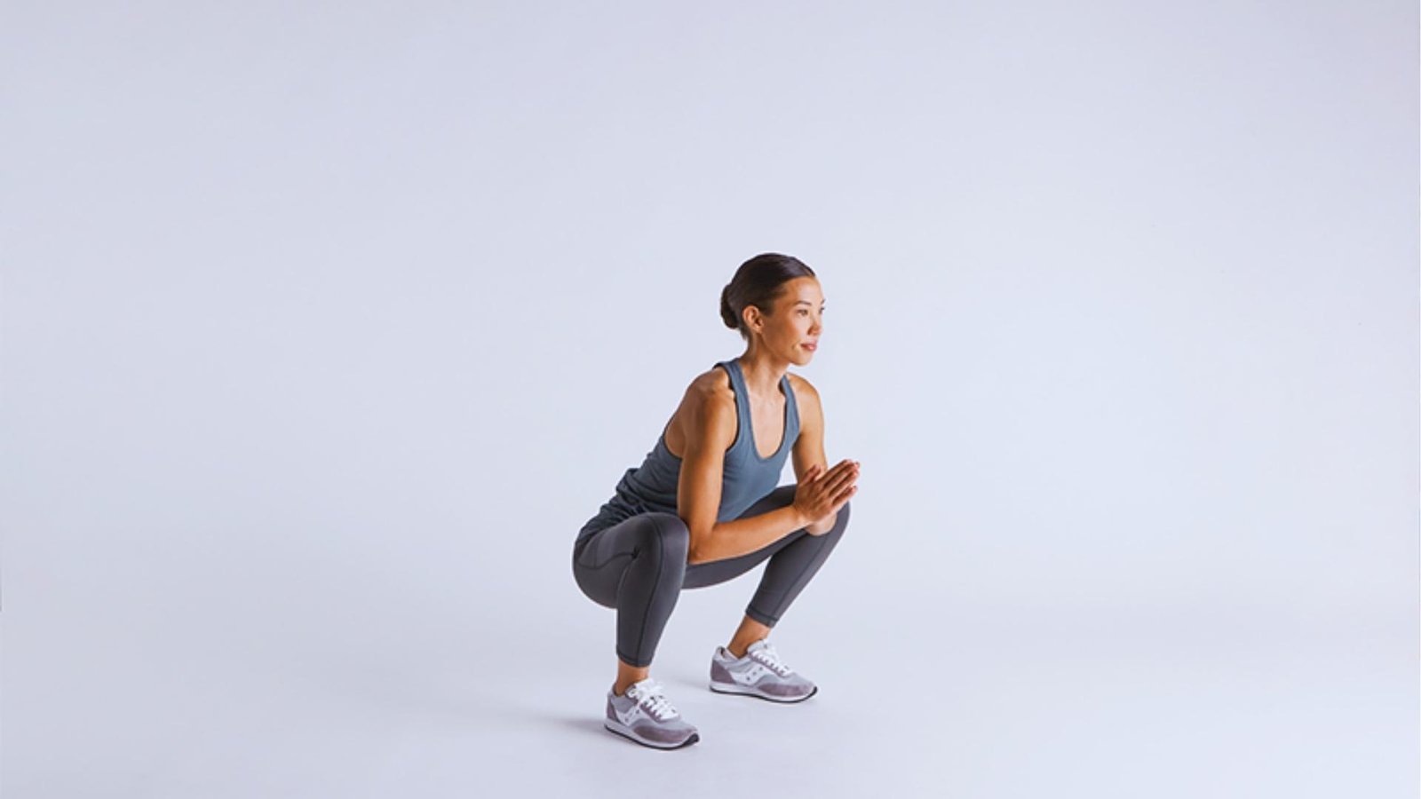 Woman in blue tank and gray leggings holds deep squat with hands in prayer, engaging pelvic floor and glutes.