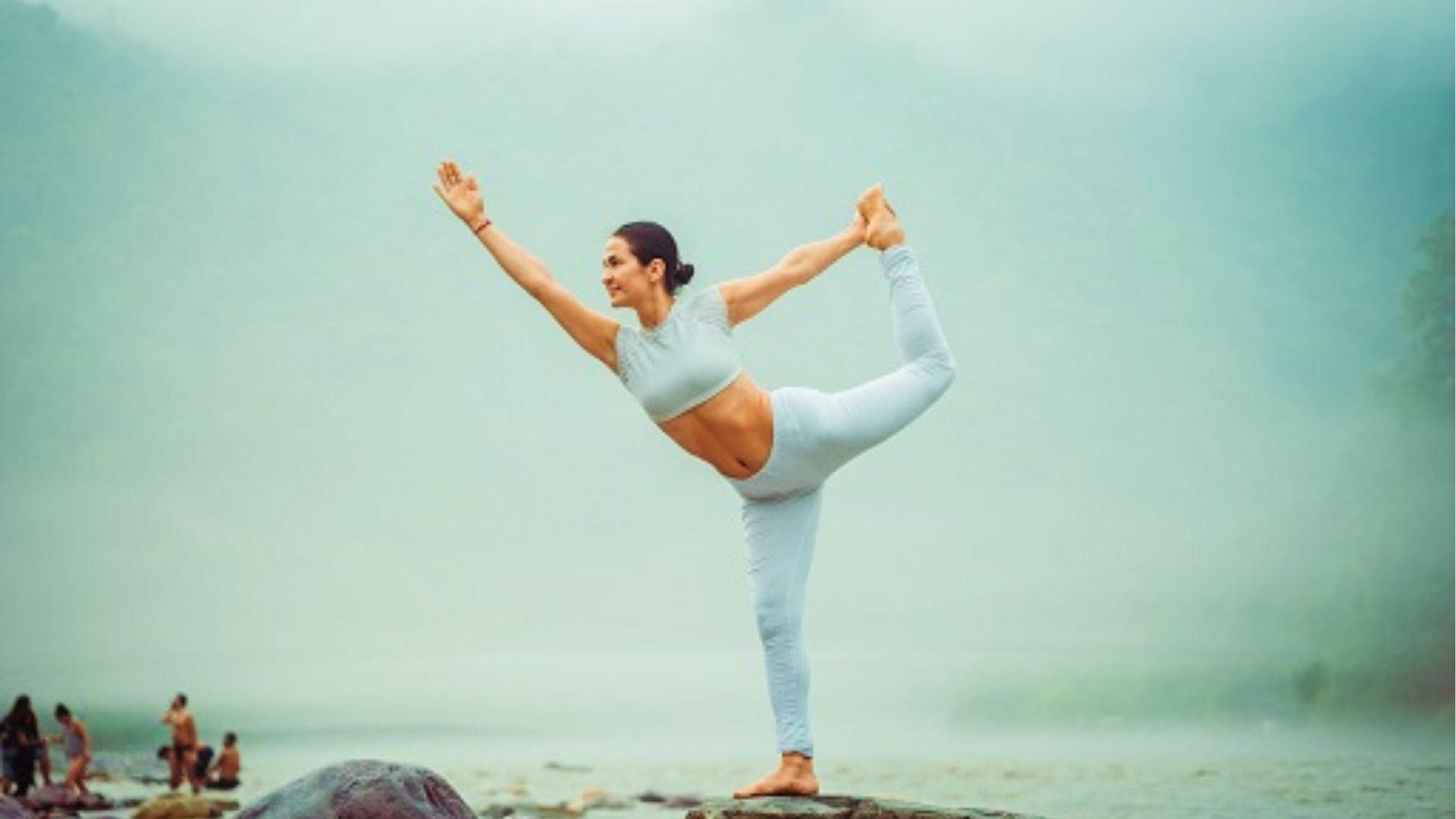 A smiling woman in white activewear holds a dancer pose on misty river rocks, people behind.