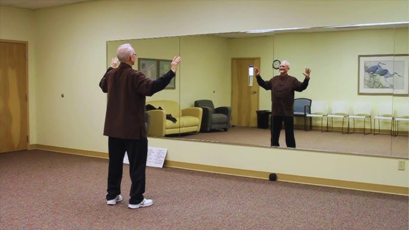 Senior man in brown tunic practices Tai Chi facing mirror in studio, hands raised, smiling at reflection.