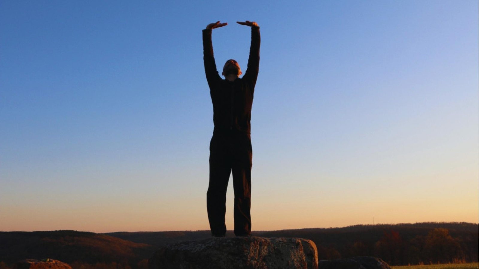 Silhouetted man in black stands on rock at sunset, arms raised overhead in Tai Chi posture against blue sky.