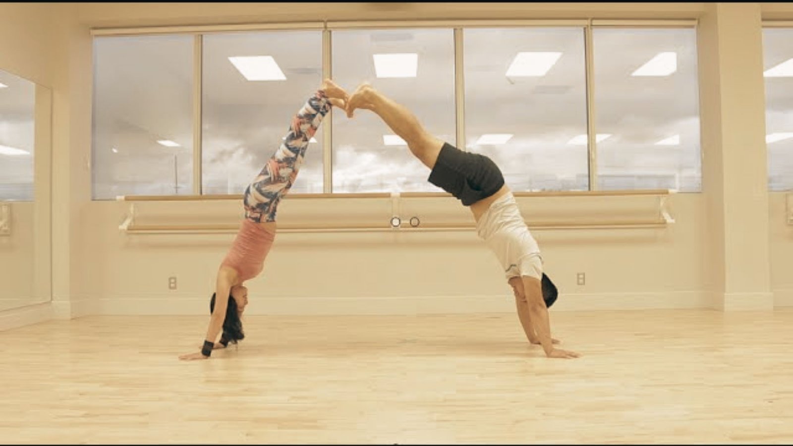 A man and woman in a yoga studio perform matching handstands facing each other, legs extended to touch at the feet.