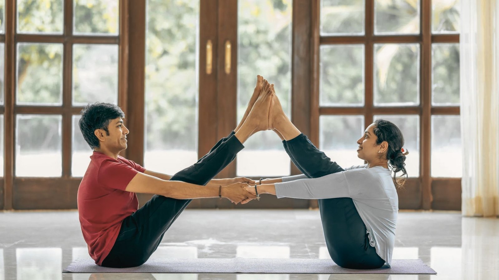 Two partners perform a supported standing forward fold in a living room, heads hanging down.