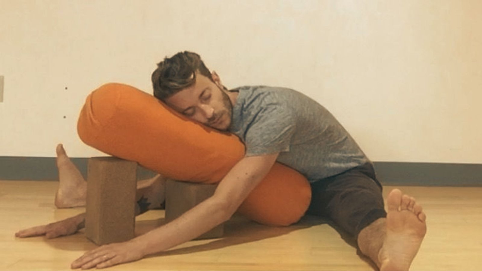 A man rests his torso on an orange bolster elevated by yoga blocks in a wide-legged forward fold.