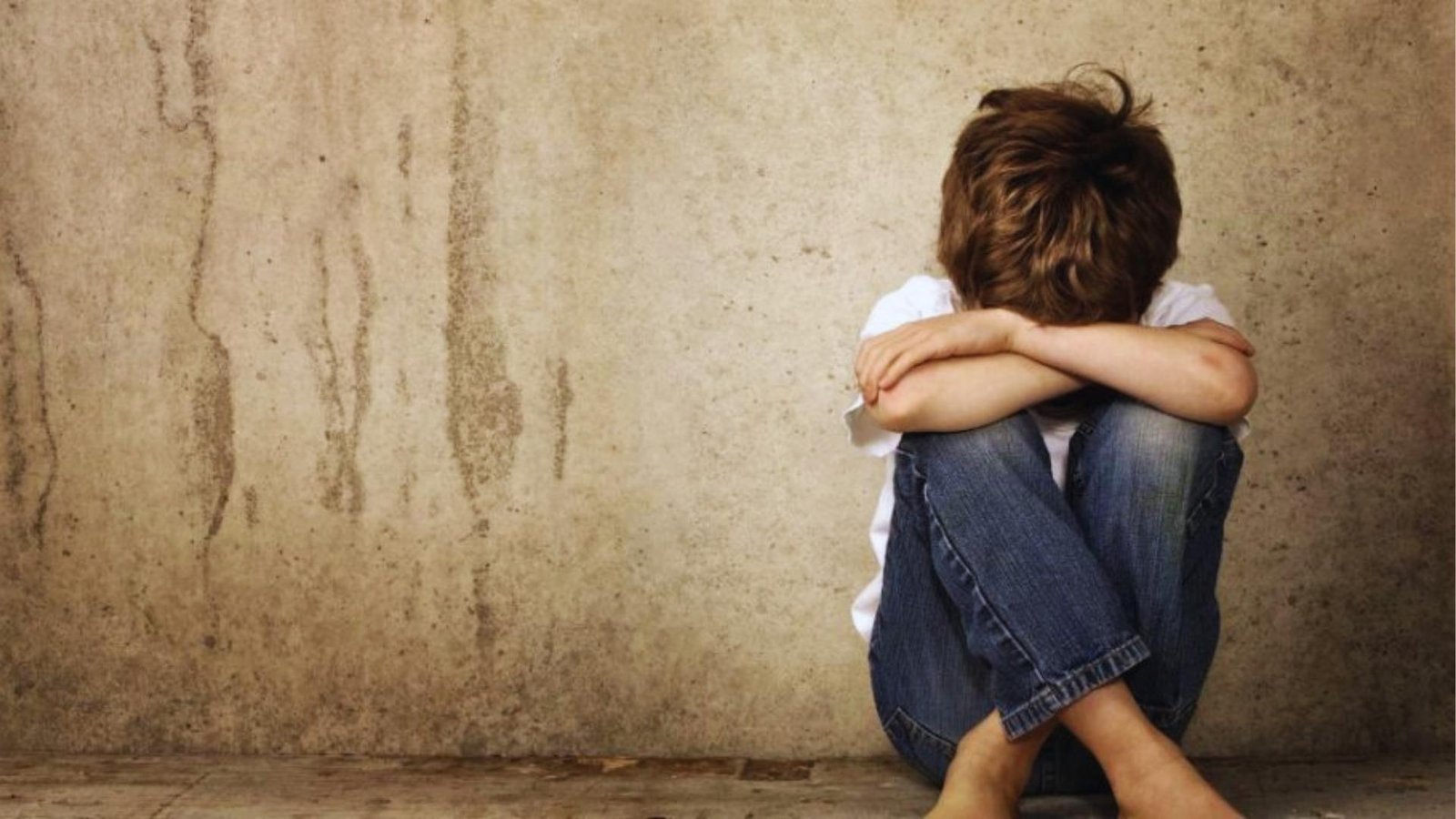 Barefoot boy in jeans and white shirt sits against concrete wall hugging knees, portraying sadness needing comfort.