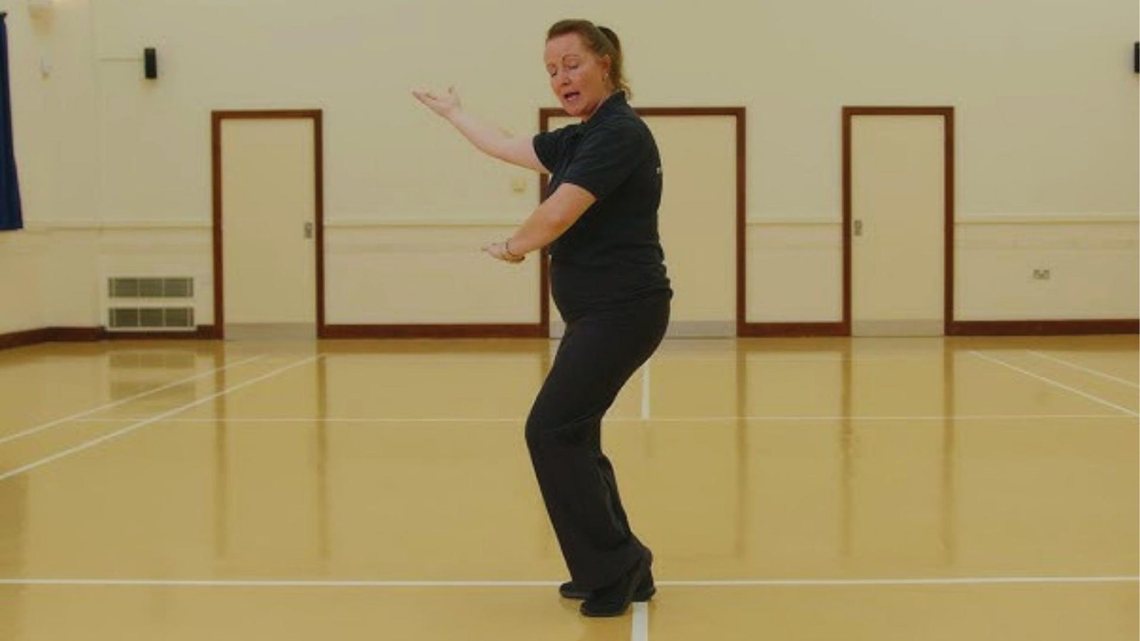 Female instructor in black demonstrates flowing Tai Chi arm movement in a bright community hall with wooden floors.
